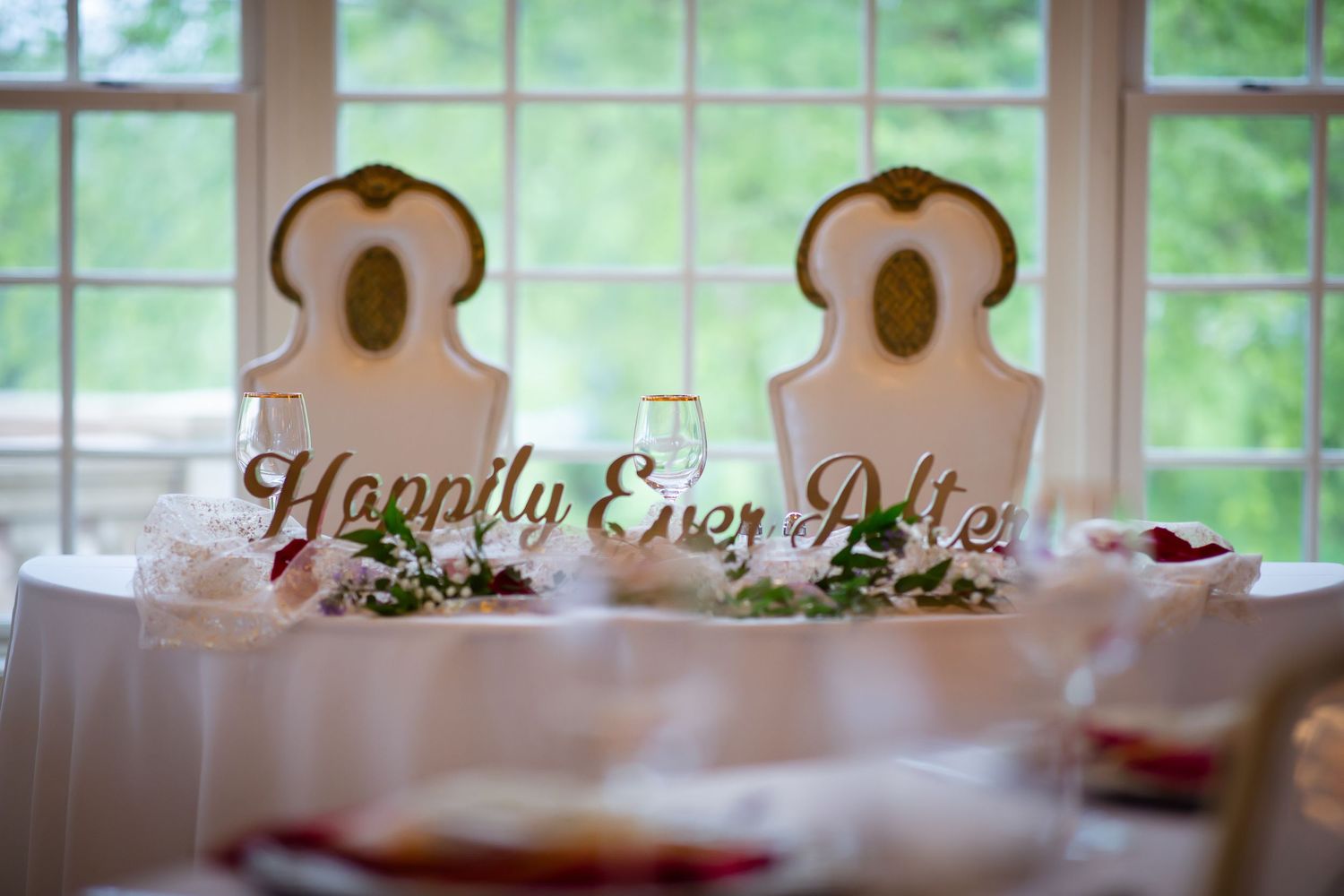 Elegant sweetheart table with ornate chairs and gold Happily Ever After sign at wedding reception.