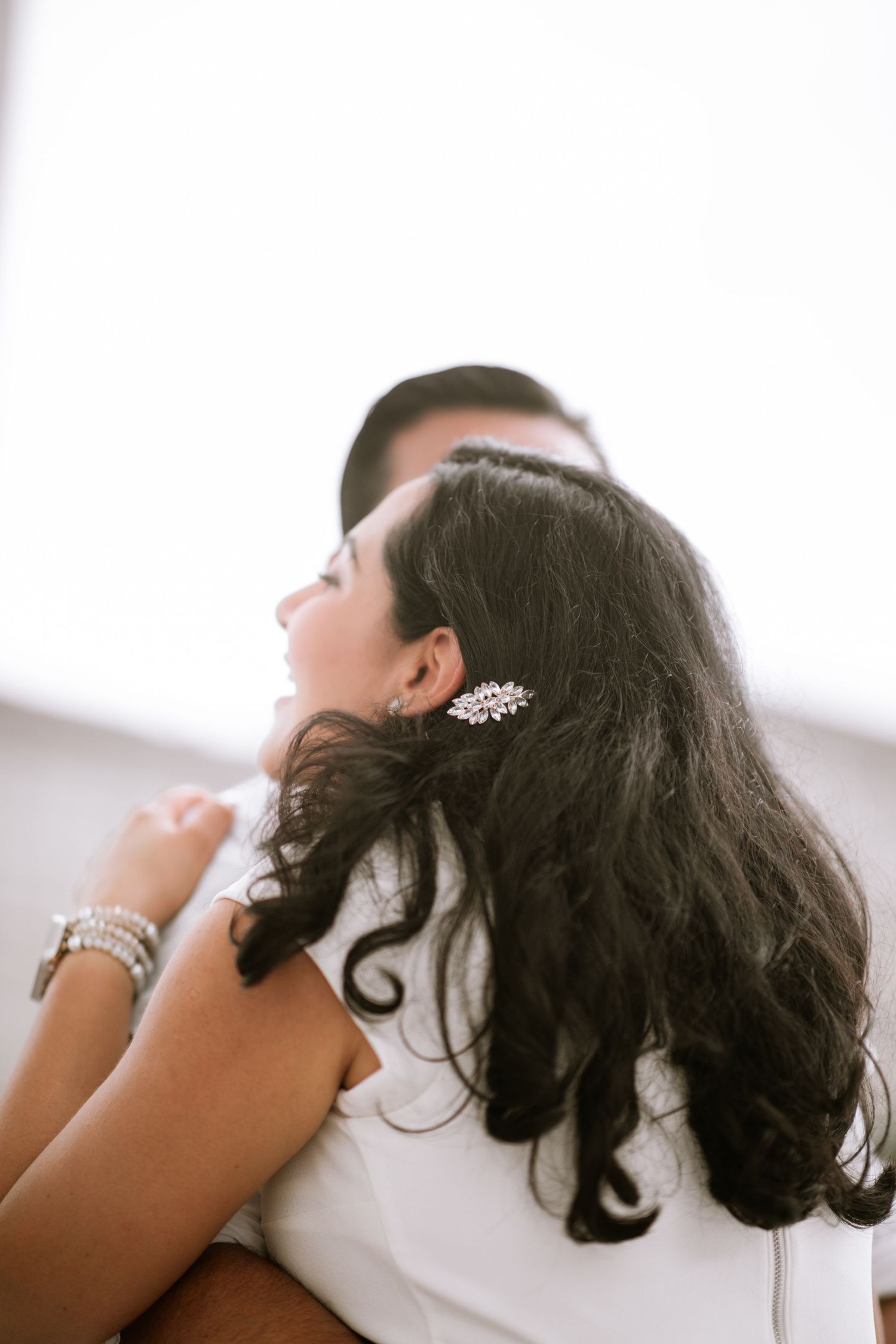 A bride with dark curly hair adorned with a crystal hair pin embraces her groom in elegant white wedding attire.