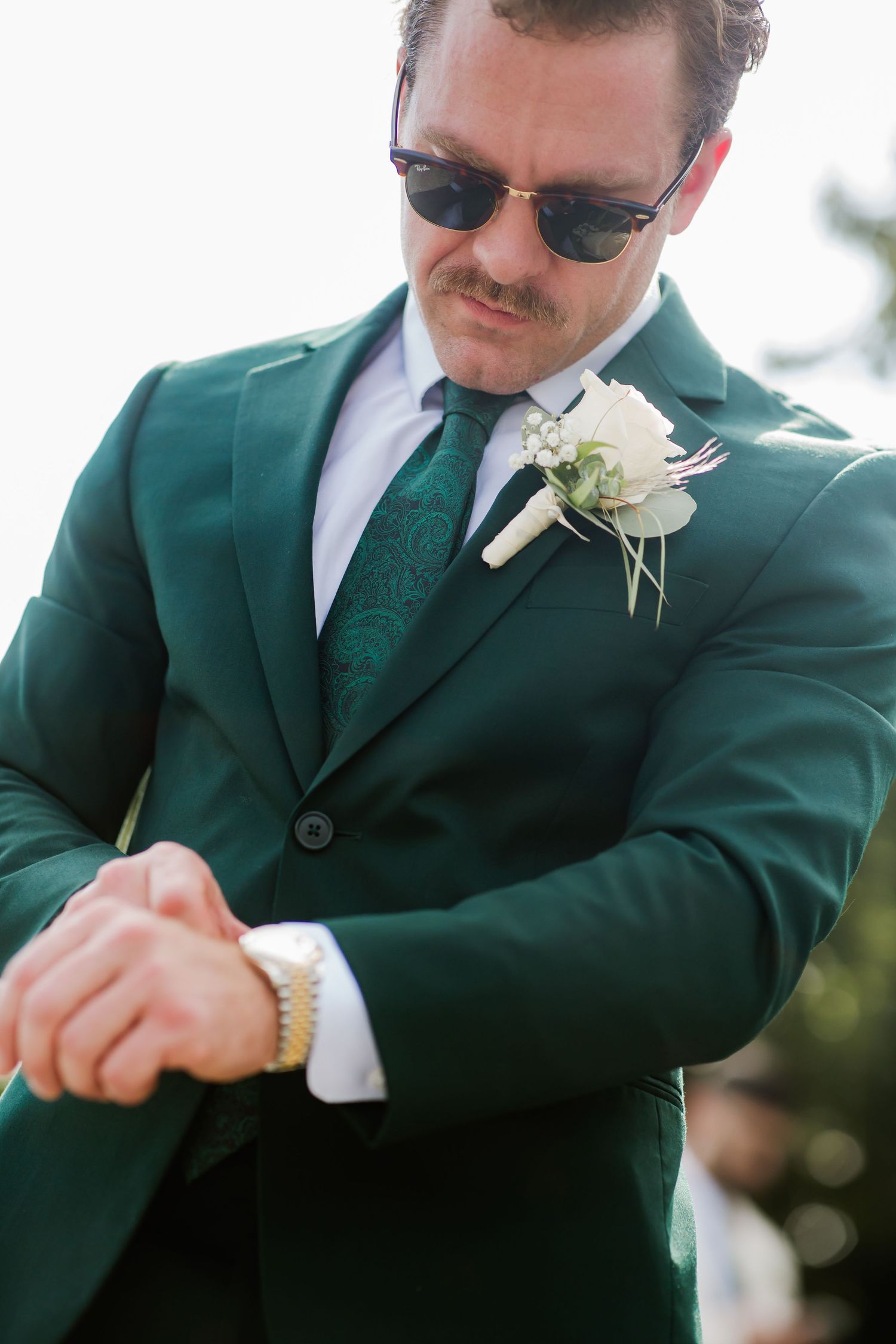 A groom in a green suit with a white boutonniere checks his watch, wearing sunglasses and a green paisley tie.