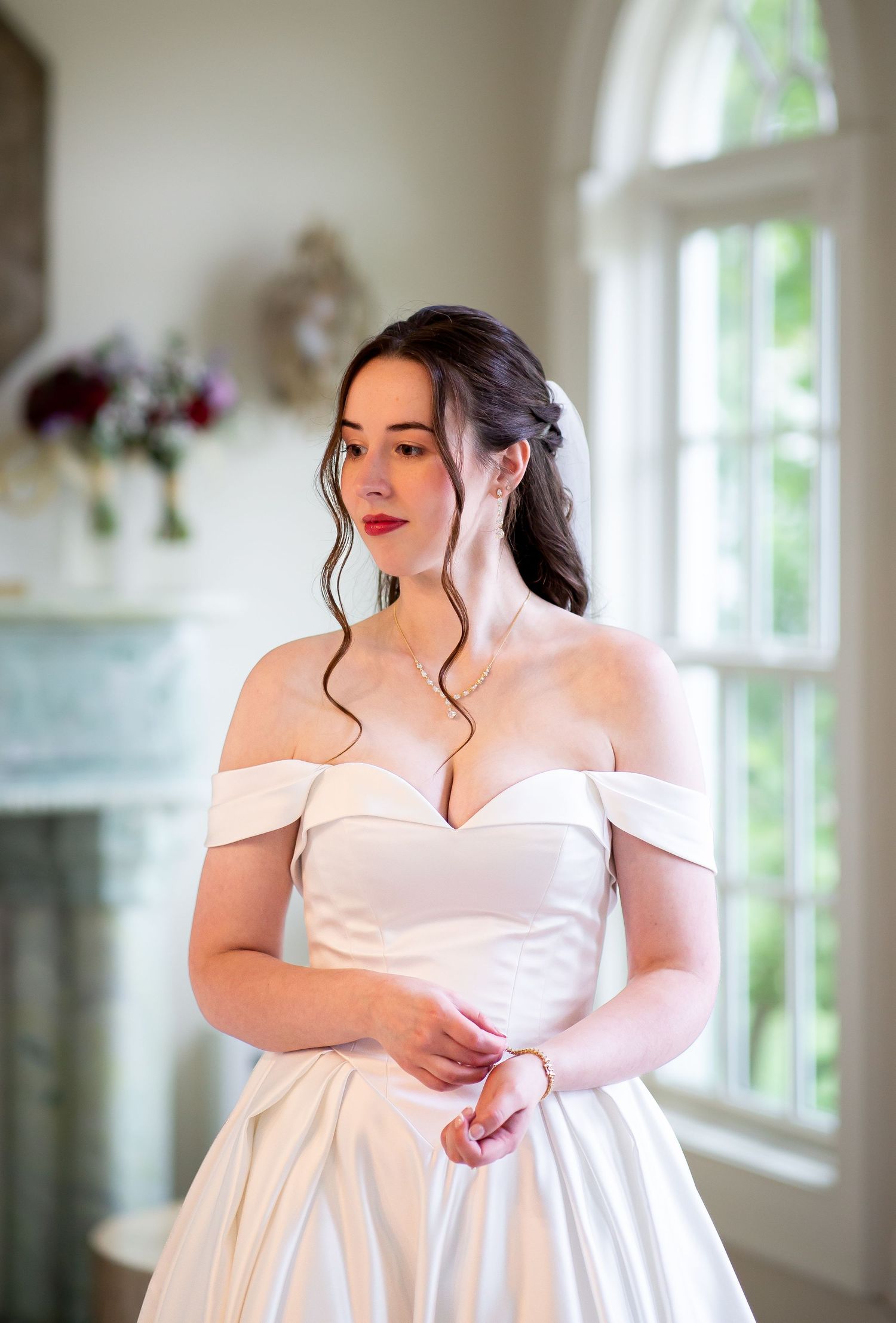 Elegant bride in off-shoulder white satin ball gown gazing out window before ceremony.