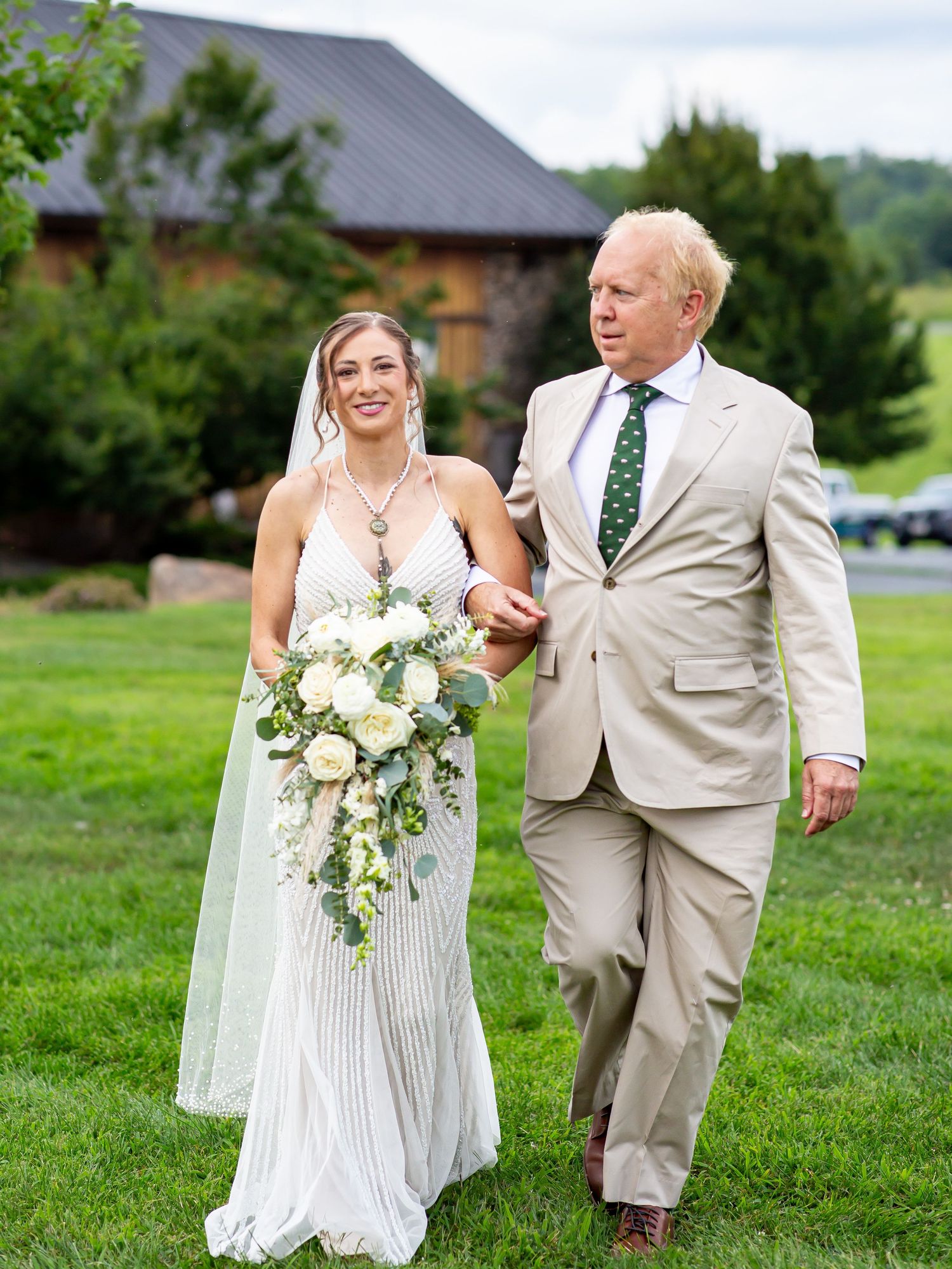 Bride in white gown with cascading bouquet walks arm-in-arm with man in tan suit on green lawn.