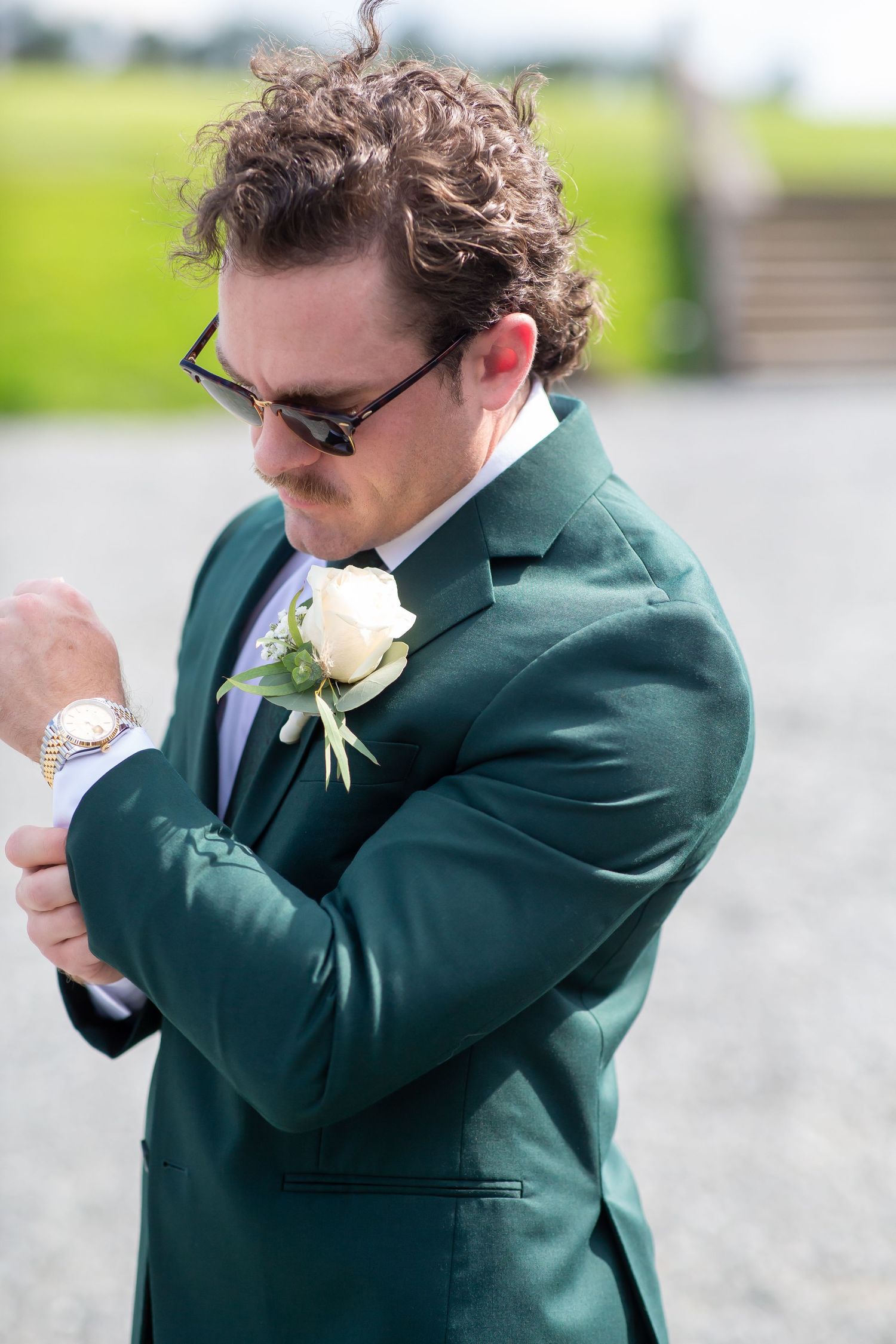 Groom in green suit with white rose boutonniere adjusting cufflinks outdoors on sunny wedding day.