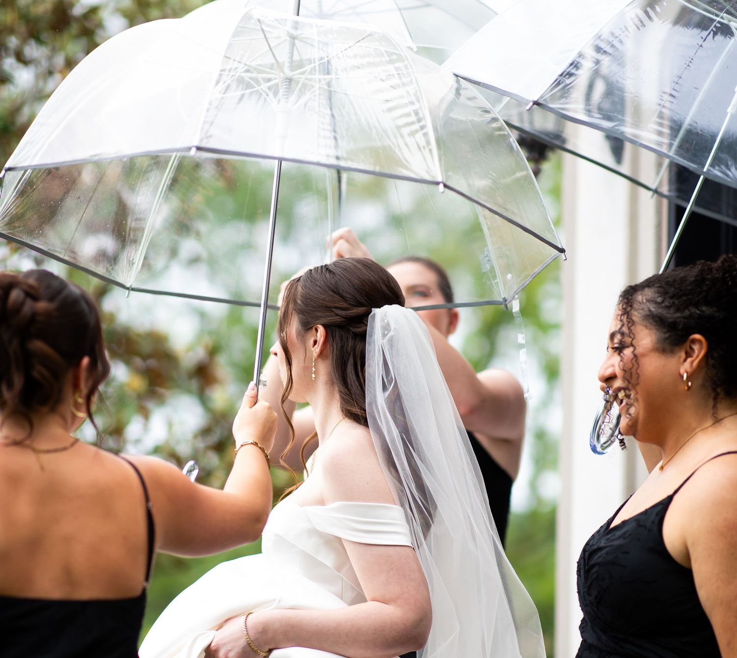 Bride in white gown and veil crying under clear umbrella surrounded by bridesmaids in black dresses outdoors.