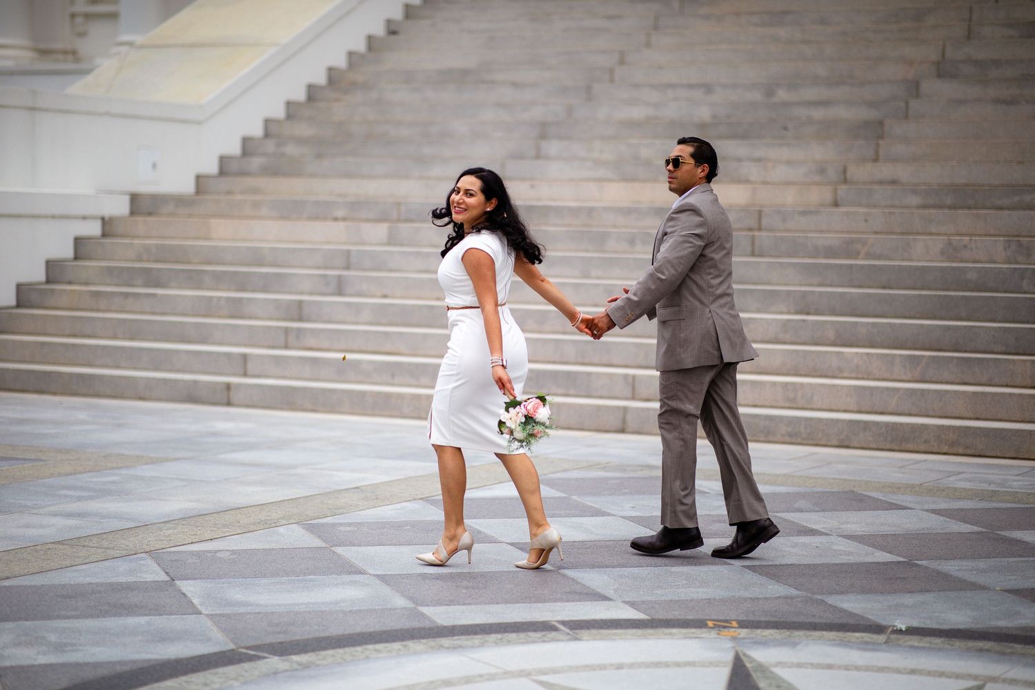 Couple in wedding attire holding hands on marble steps, bride in white dress with bouquet, groom in gray suit.