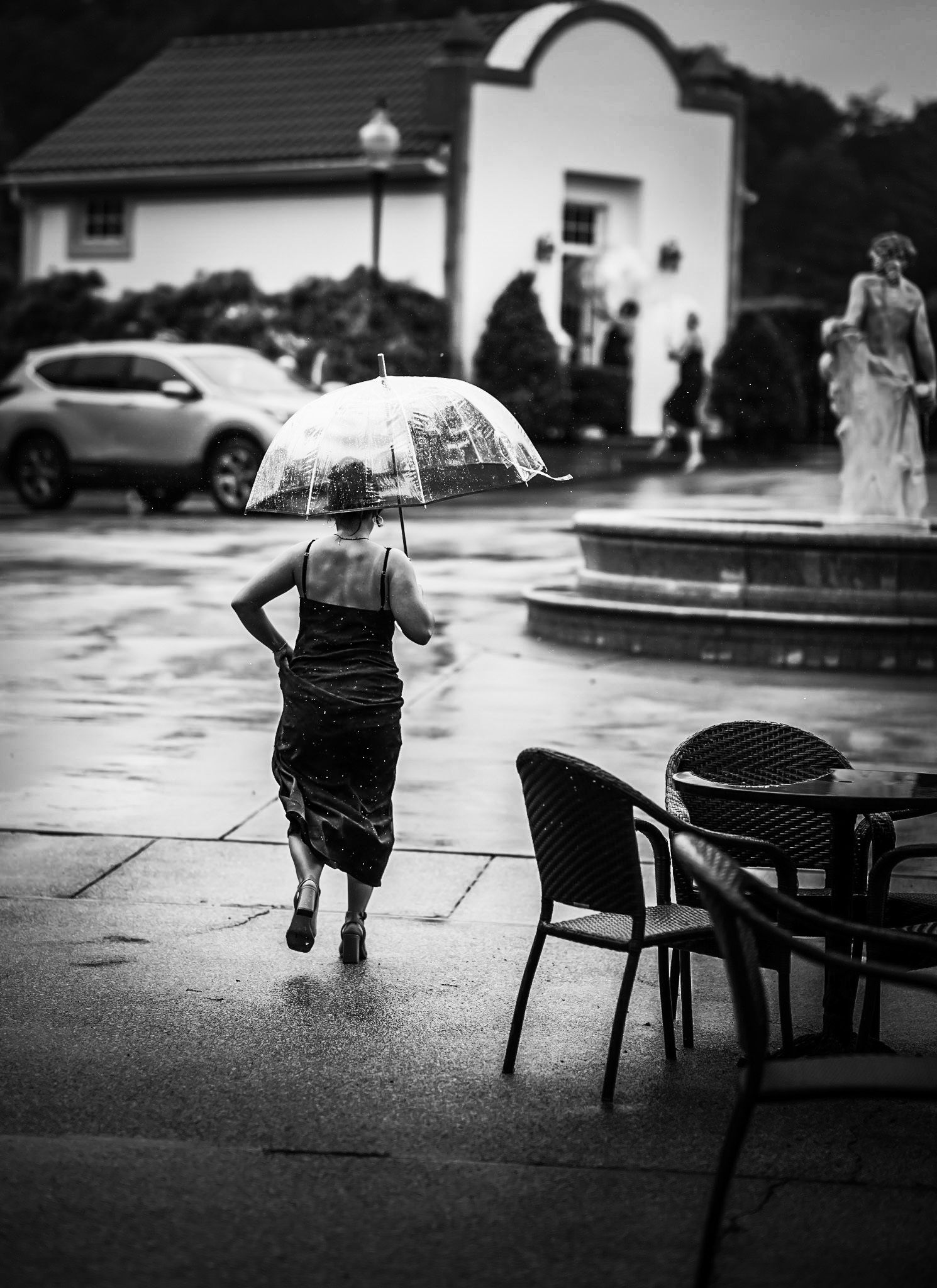 Woman in black dress running with clear umbrella in rain near fountain at wedding venue, black and white.