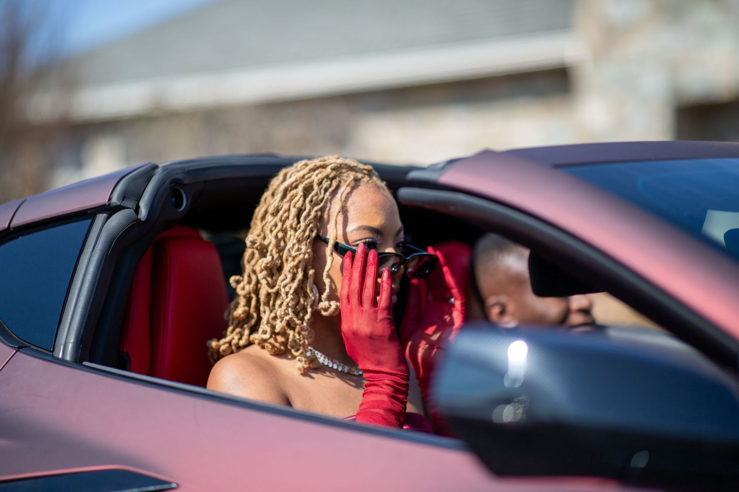 Glamorous woman in red gloves and sunglasses posing inside a red convertible with a man in background.