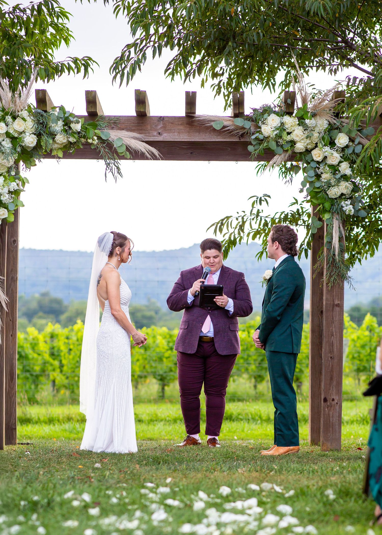 Outdoor wedding ceremony under a wooden pergola with floral arrangements, bride in white, groom in teal, officiant in purple.