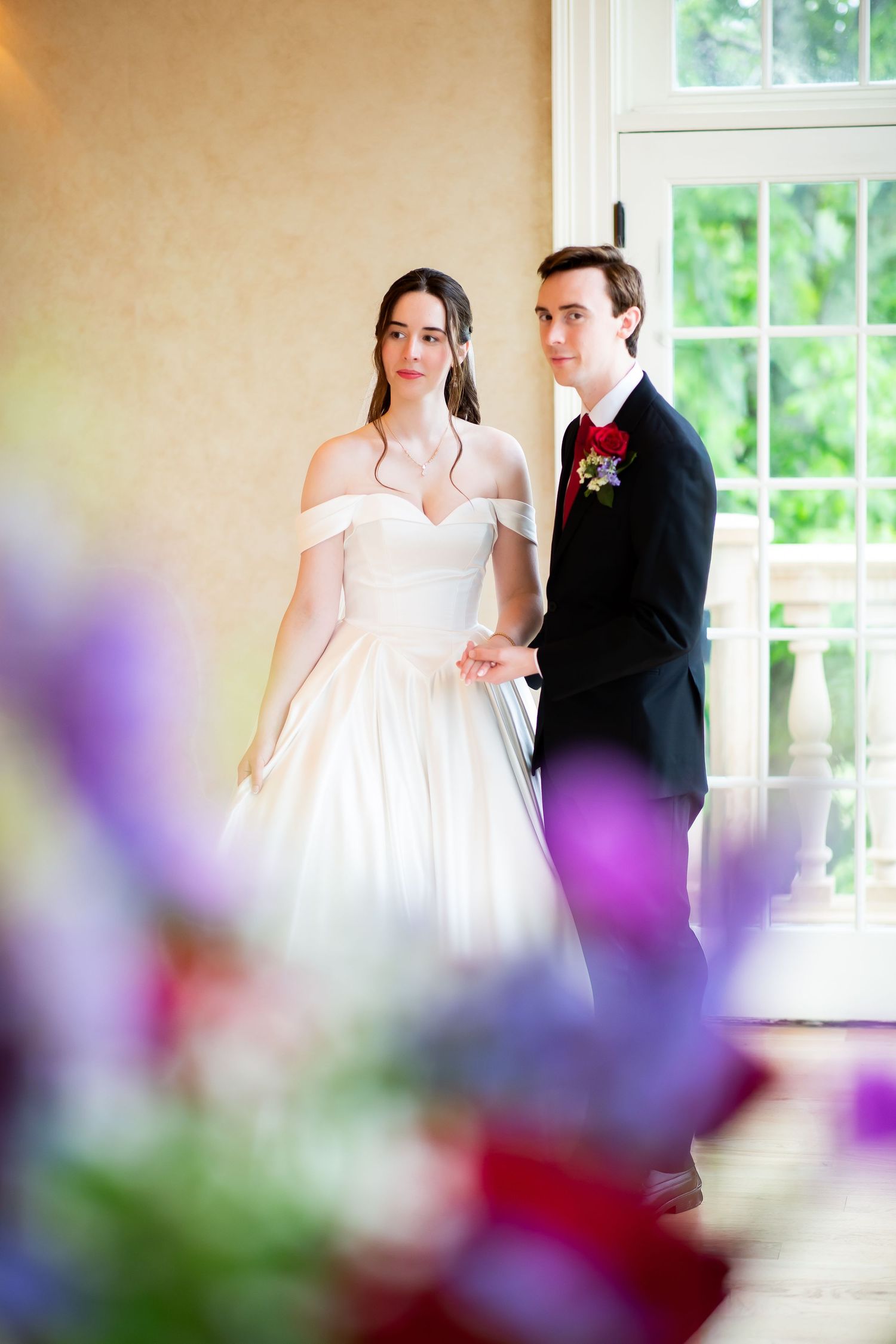 Bride in off-shoulder white gown and groom in black suit holding hands near sunlit window.