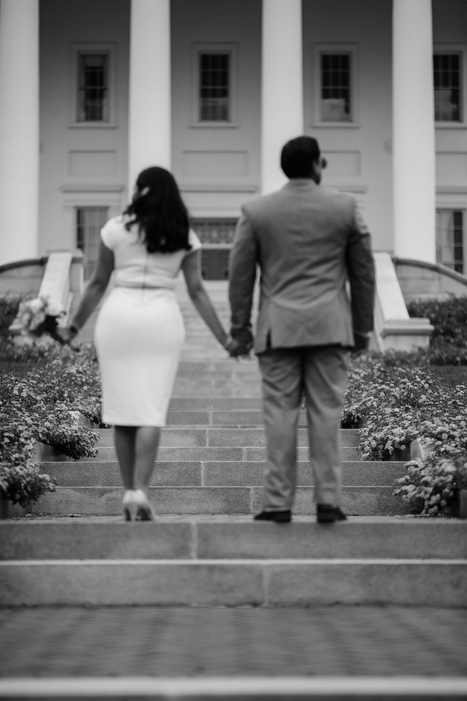 Black and white photo of a couple holding hands walking up steps toward a grand white building.