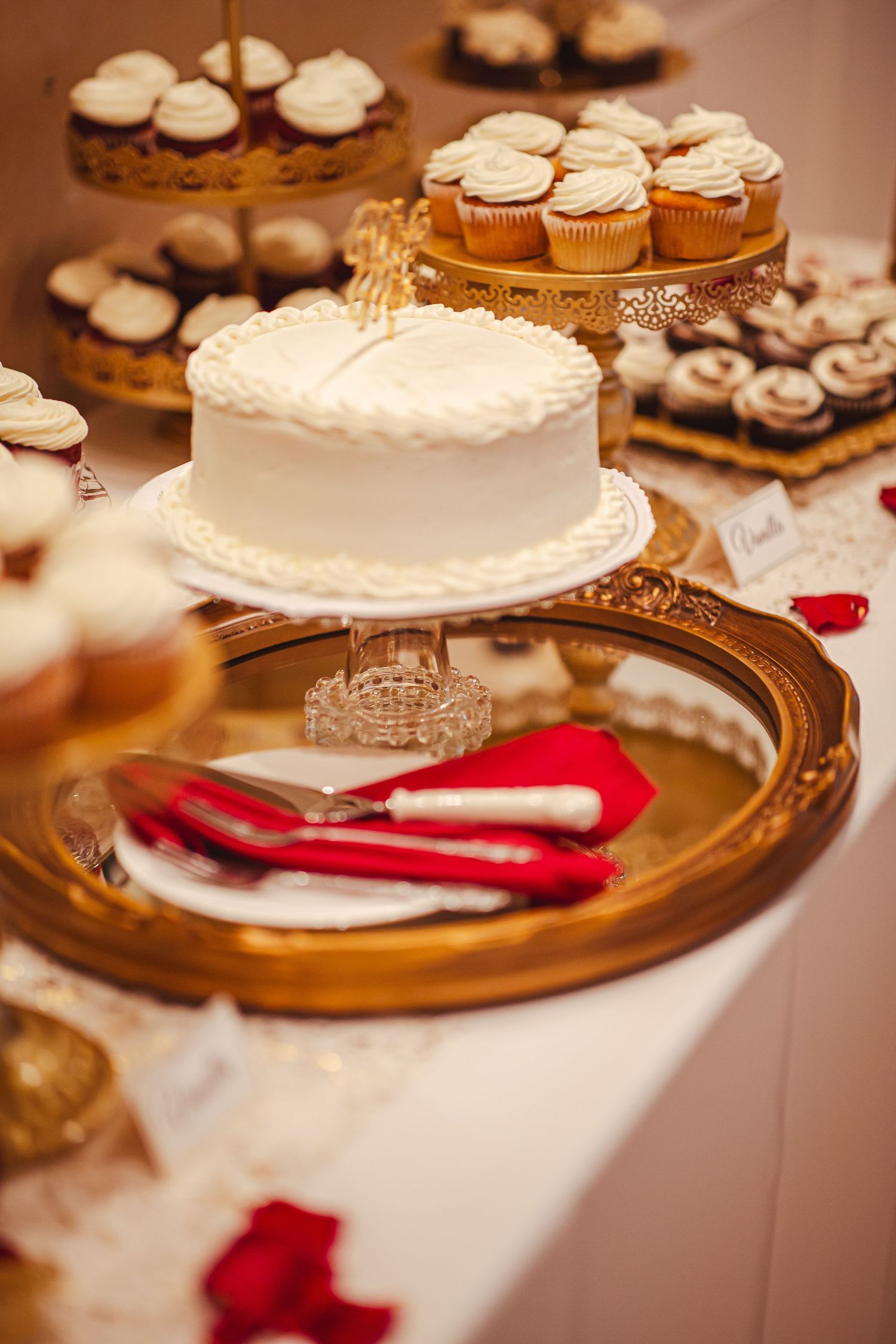 White wedding cake surrounded by cupcakes on gold stands with red napkins on dessert table.