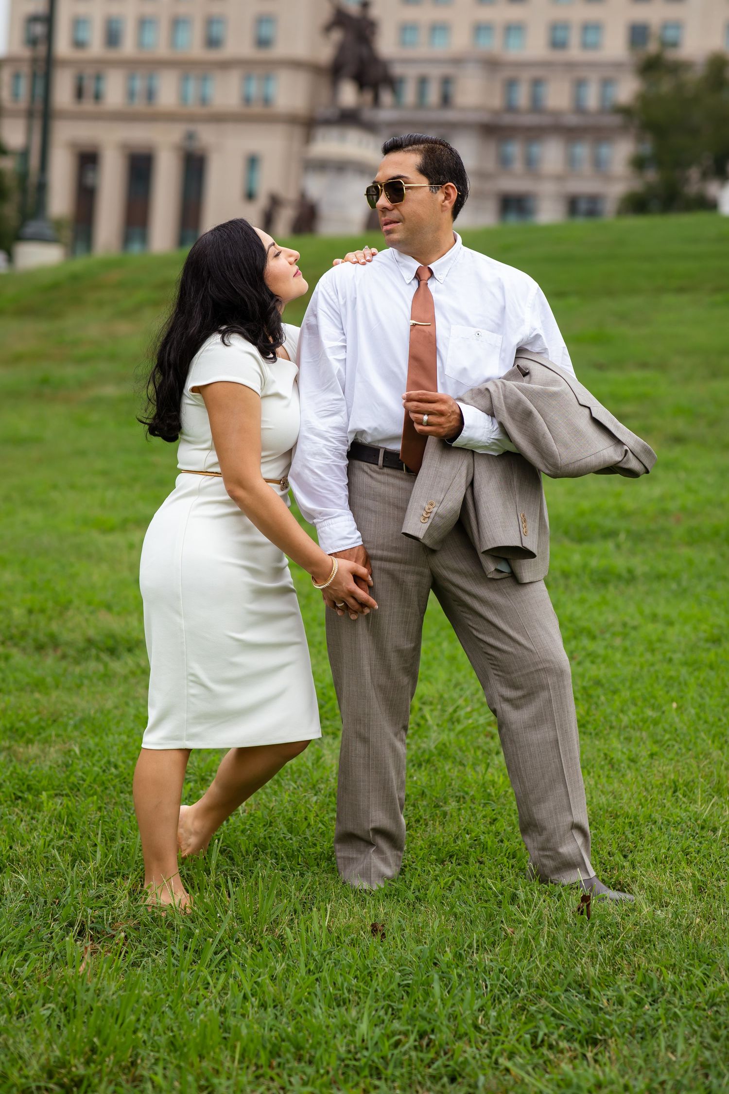 Couple holding hands on green lawn near historic building, woman in white dress, man in suit holding jacket.
