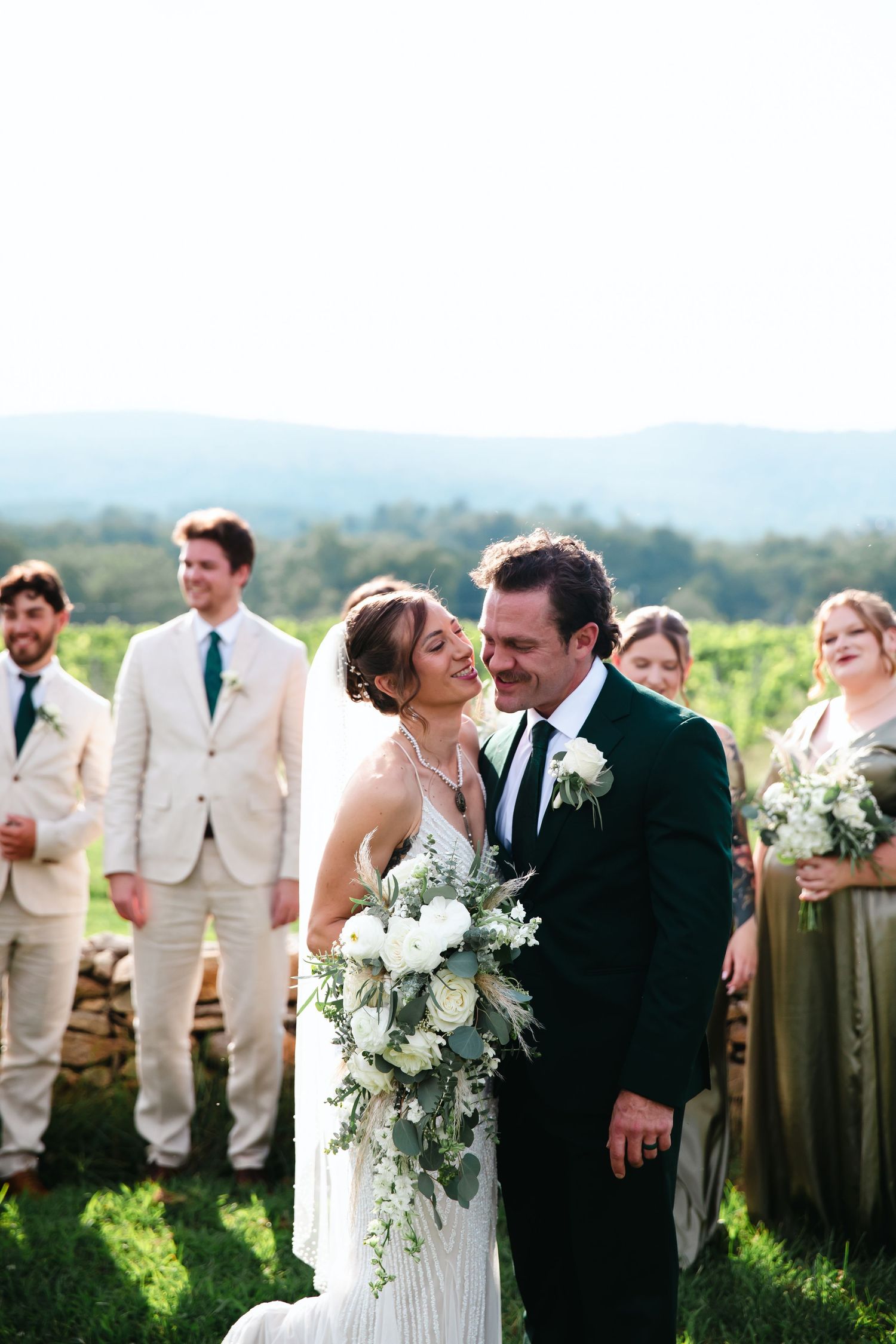 Bride and groom share a romantic moment with wedding party and vineyard hills backdrop on their wedding day.