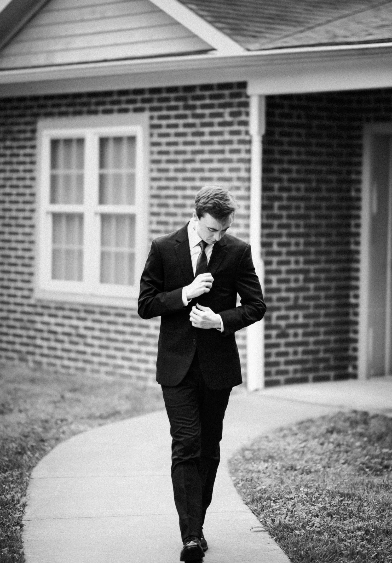 Groom in black suit adjusting his tie while walking along a brick building pathway in black and white.
