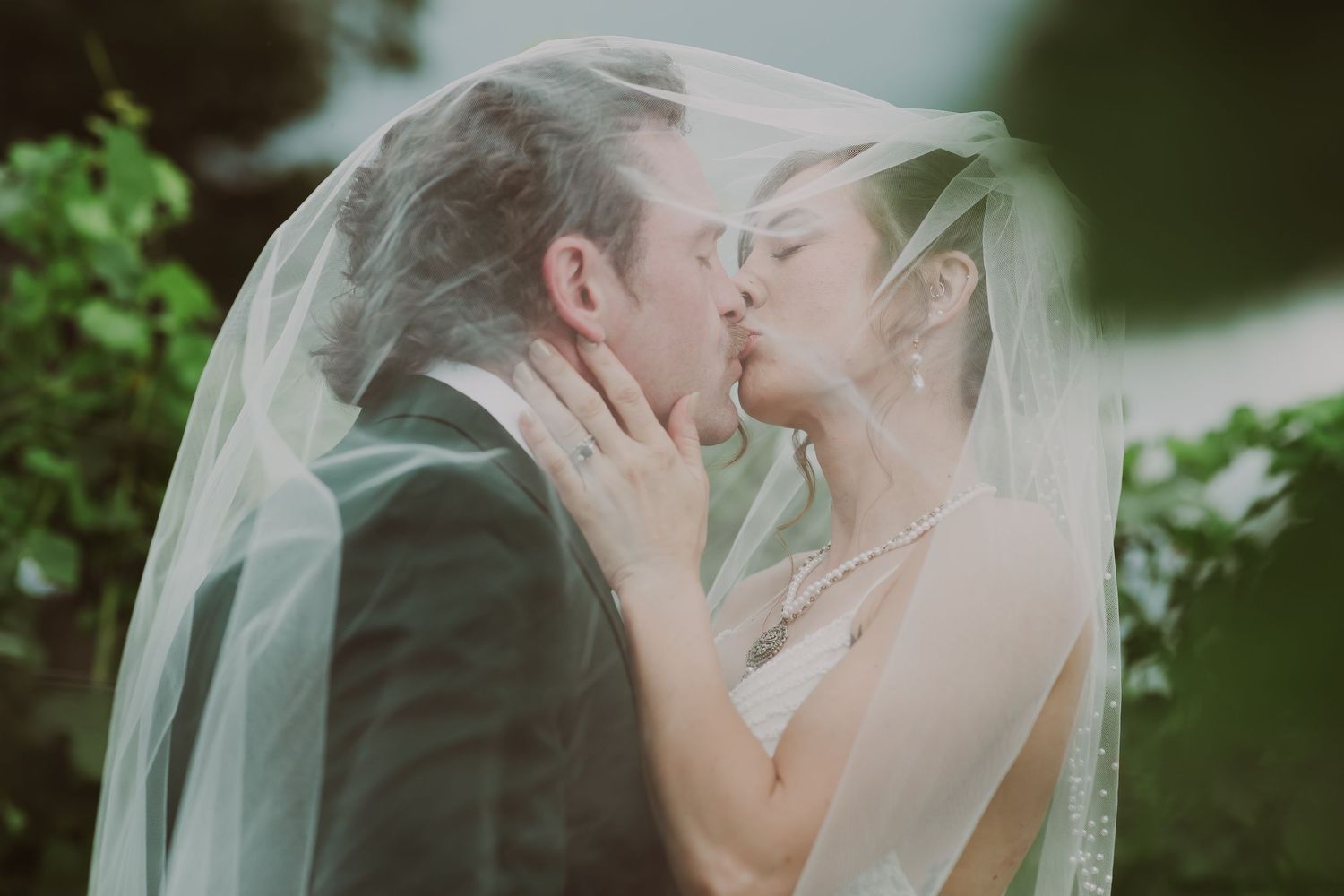 Bride and groom sharing a romantic kiss under a flowing white wedding veil outdoors.