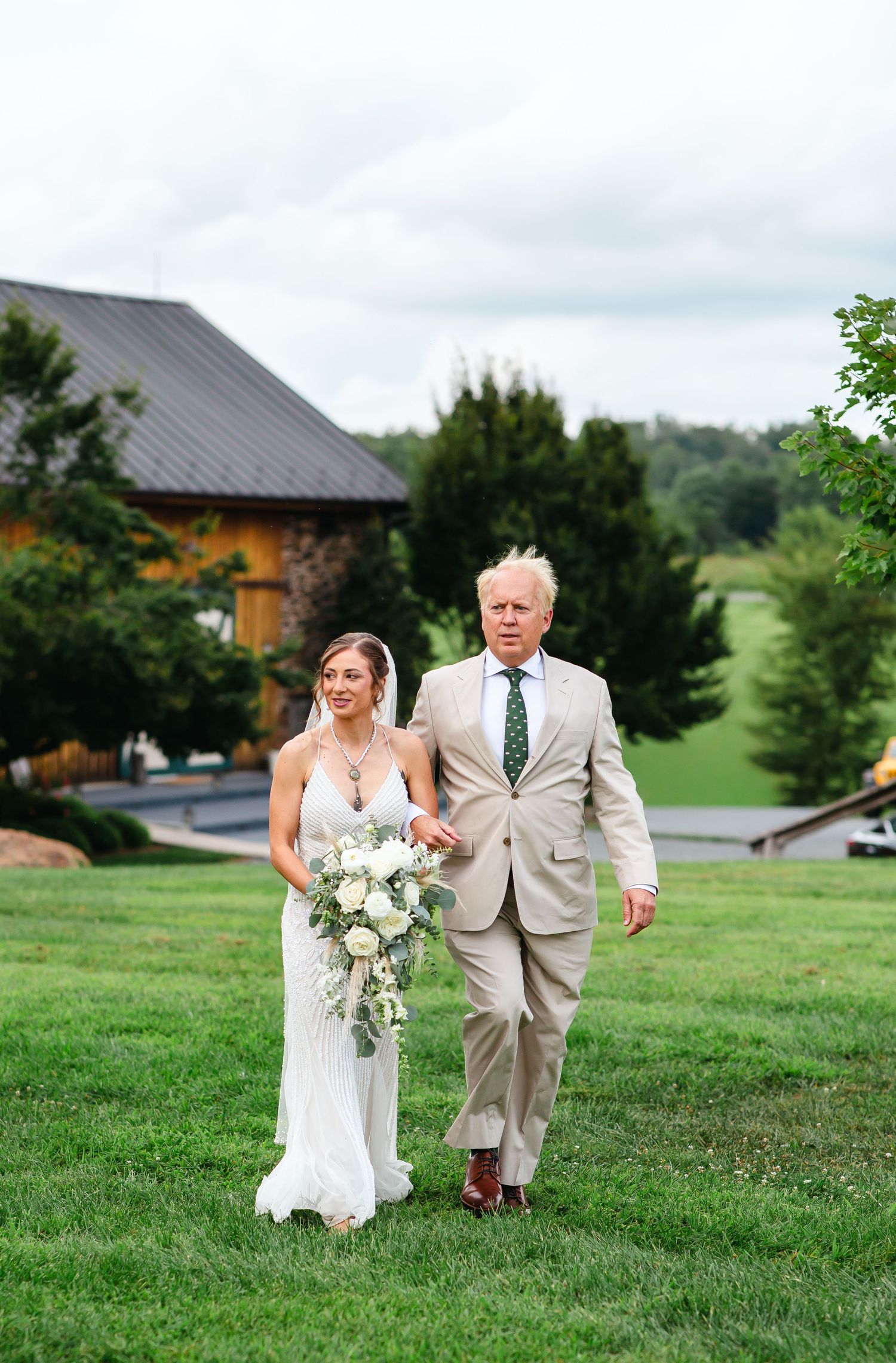 Bride in white gown holding bouquet walks with older man in tan suit on green lawn at outdoor wedding venue.