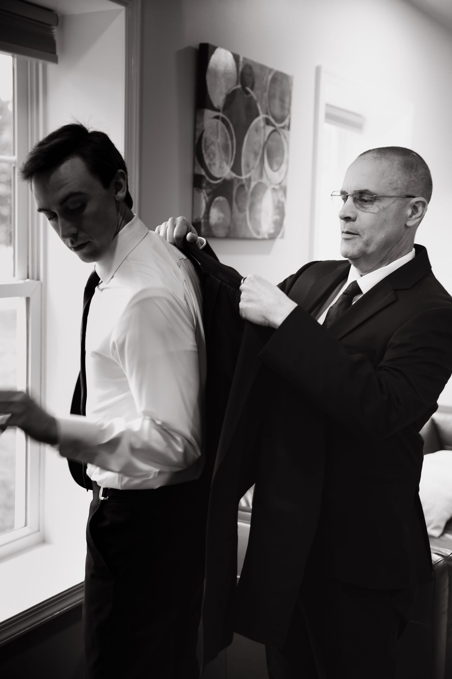 Older man helping young groom put on suit jacket while getting ready for wedding ceremony.