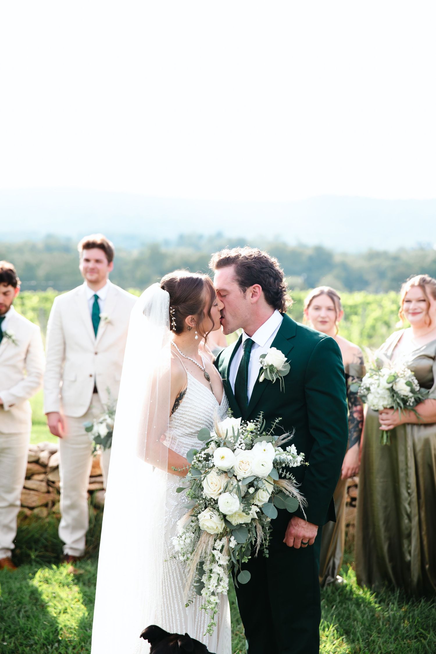 Bride and groom share first kiss at outdoor vineyard wedding ceremony with bridal party in green and ivory attire.