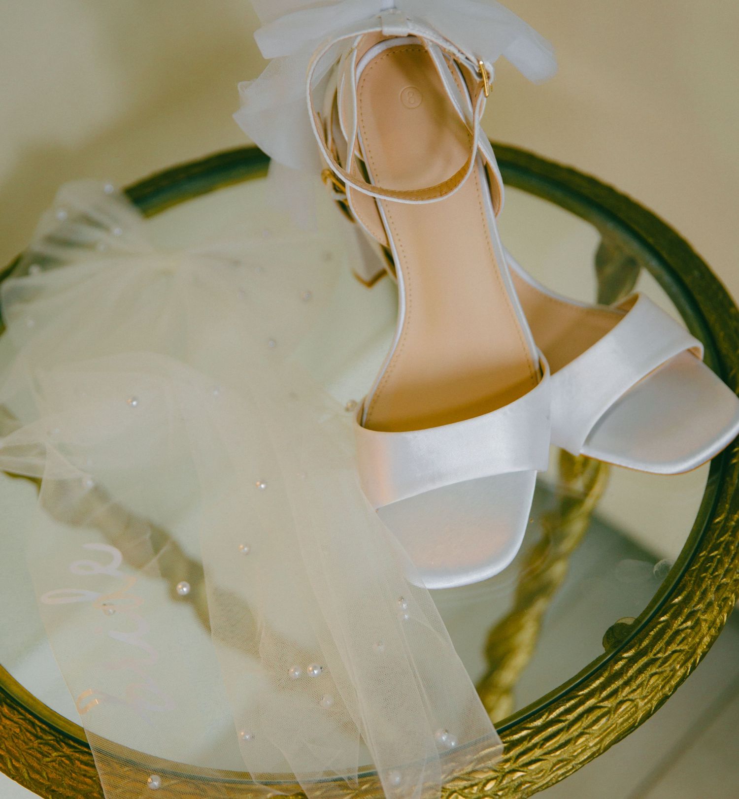 Elegant white block-heel bridal sandals resting on a mirrored tray with a pearl-trimmed wedding veil.