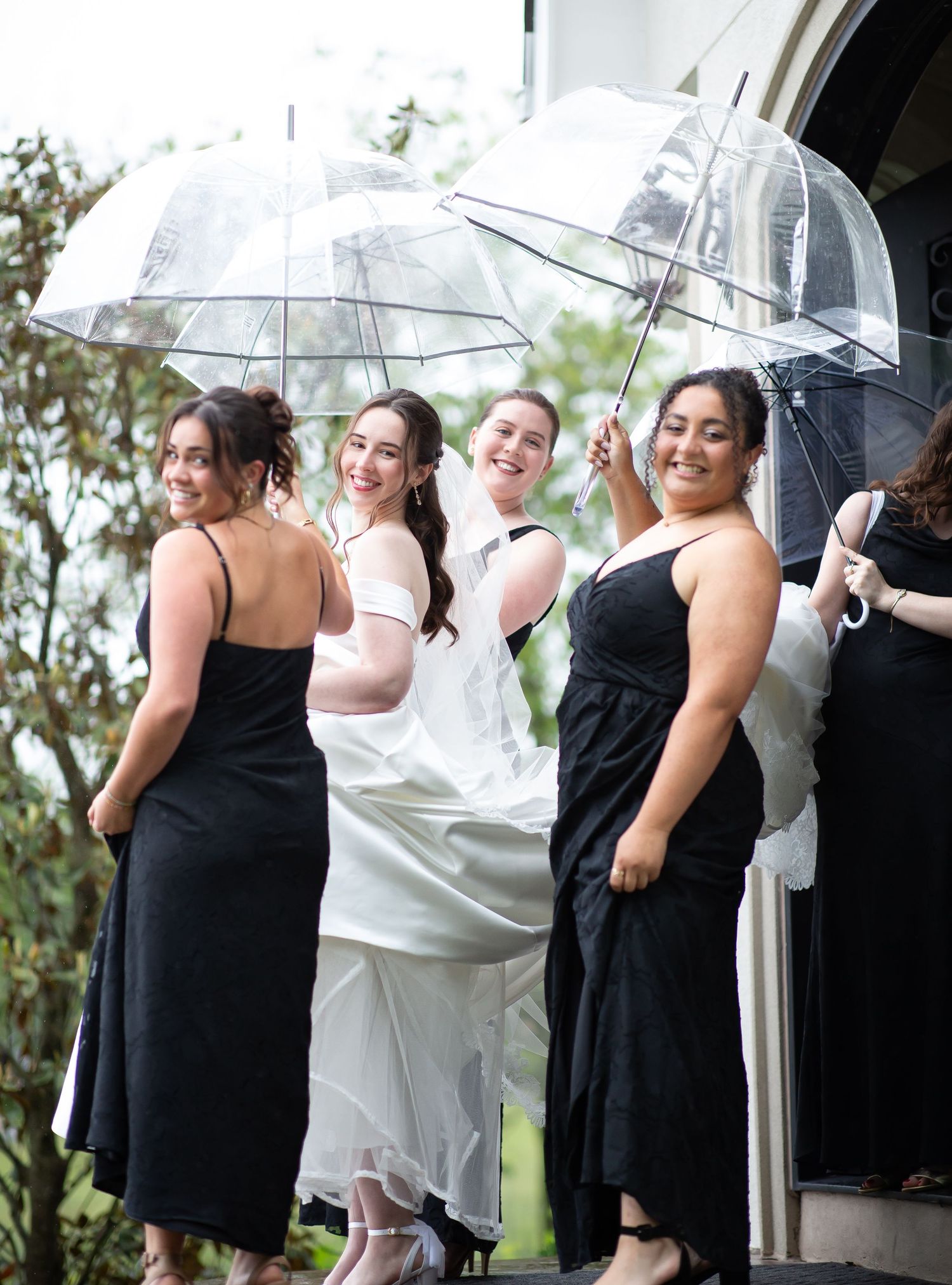 Smiling bride in white veil with bridesmaids in black dresses holding clear umbrellas on rainy wedding day.