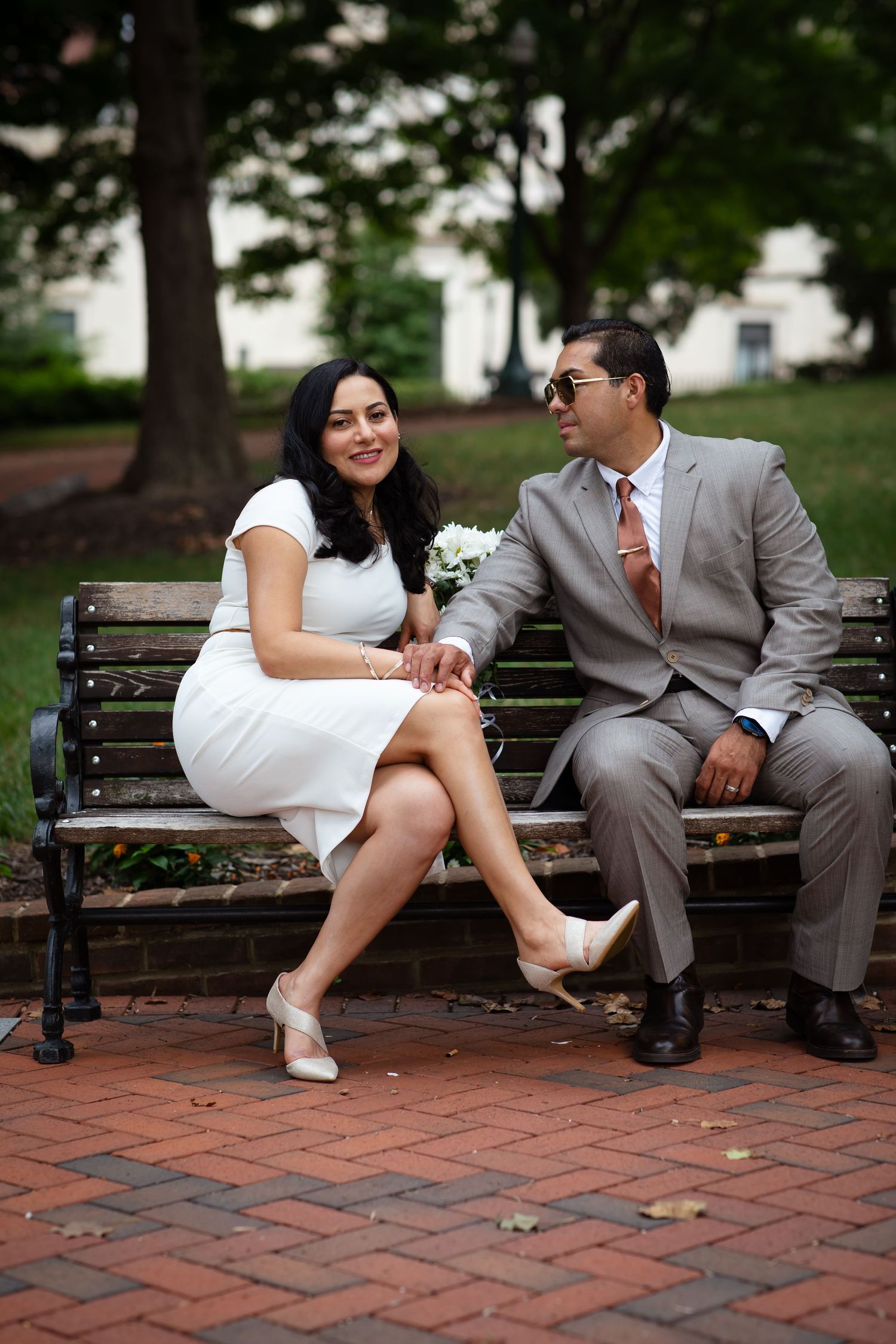 A couple in wedding attire sits on a park bench, woman in white dress holding bouquet, man in gray suit with sunglasses.