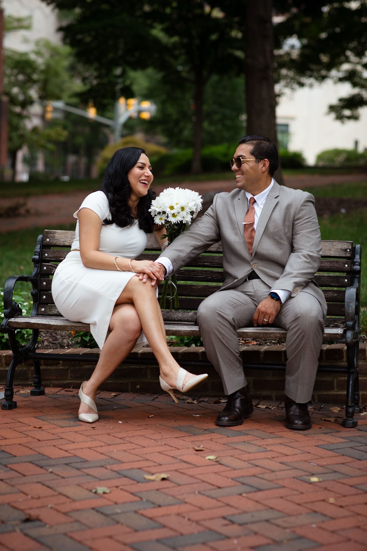 Smiling couple in wedding attire sitting on a park bench, bride holding white bouquet, groom in gray suit with pink tie.