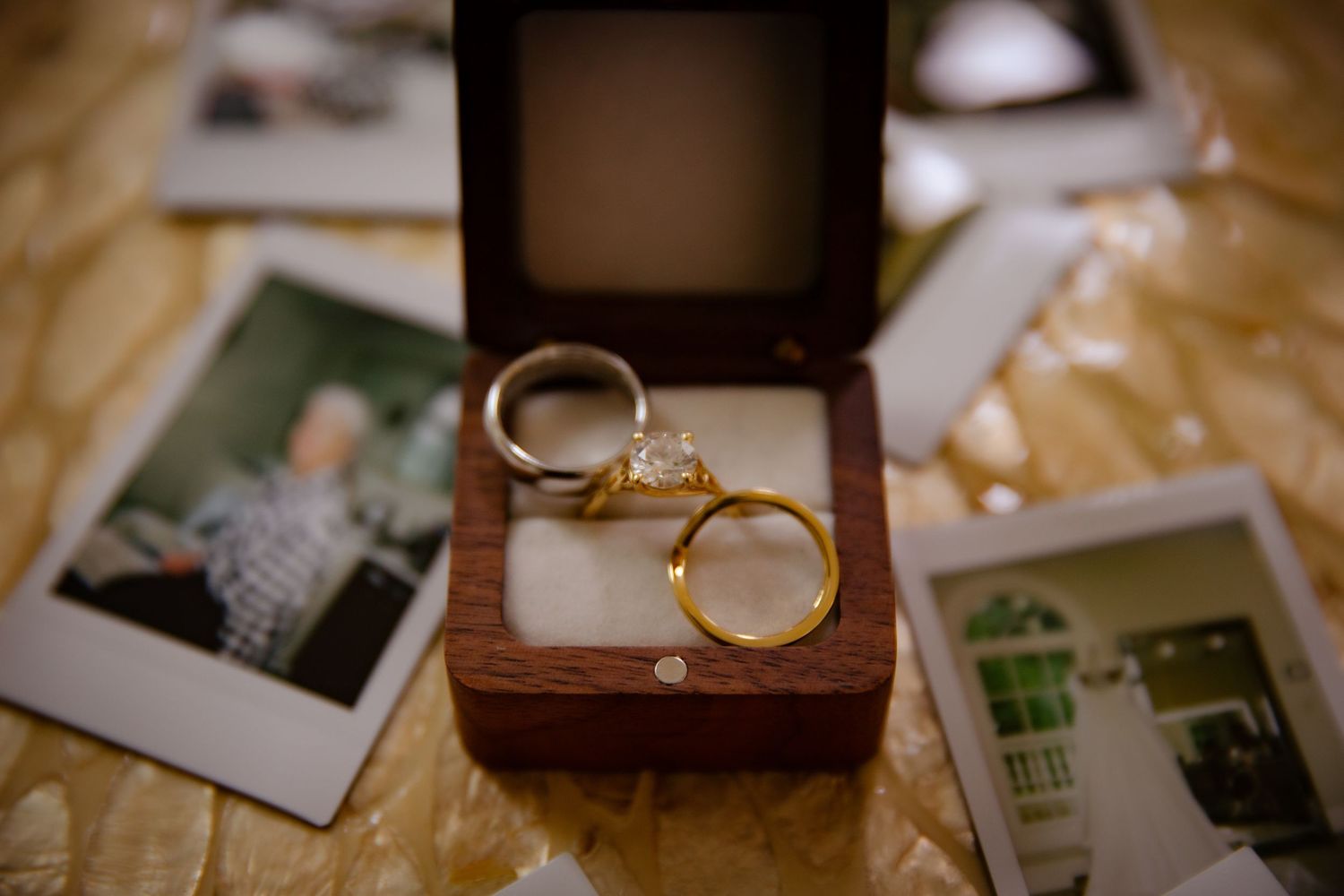 Gold diamond engagement ring and wedding bands displayed in a wooden ring box with Polaroid photos.