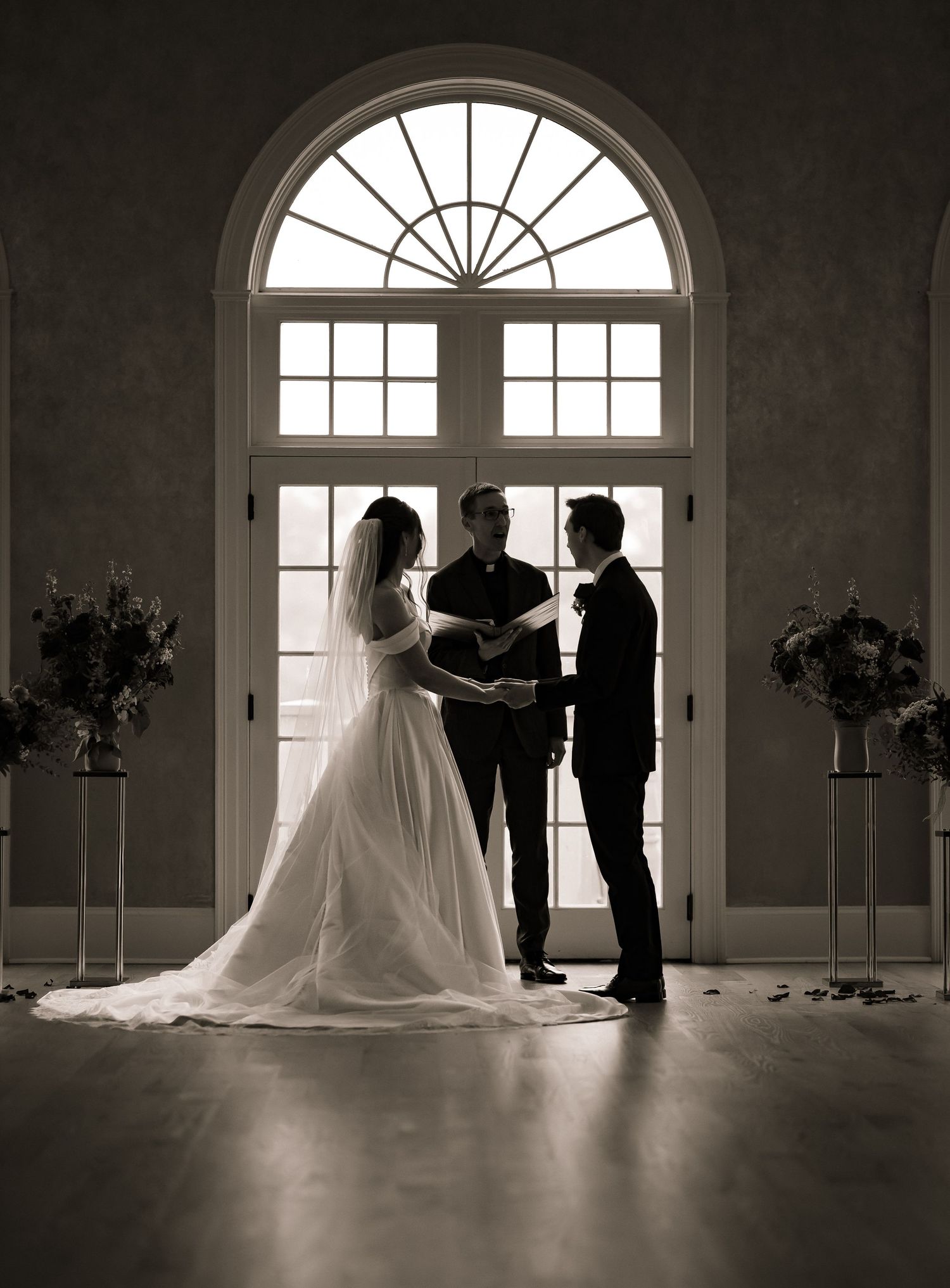 Silhouette of a bride and groom exchanging vows before an arched window in a black and white photo.