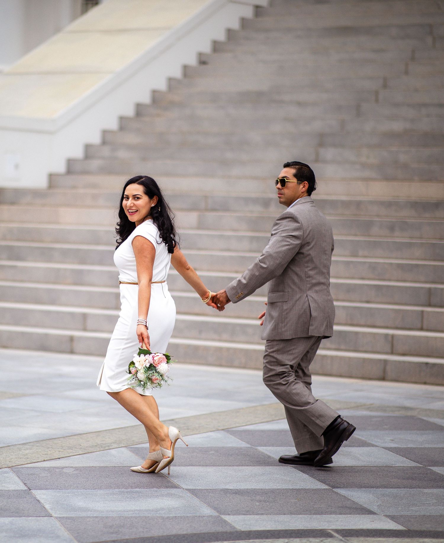 Bride in white dress holding bouquet walks hand-in-hand with groom in gray suit on checkered plaza near stairs.