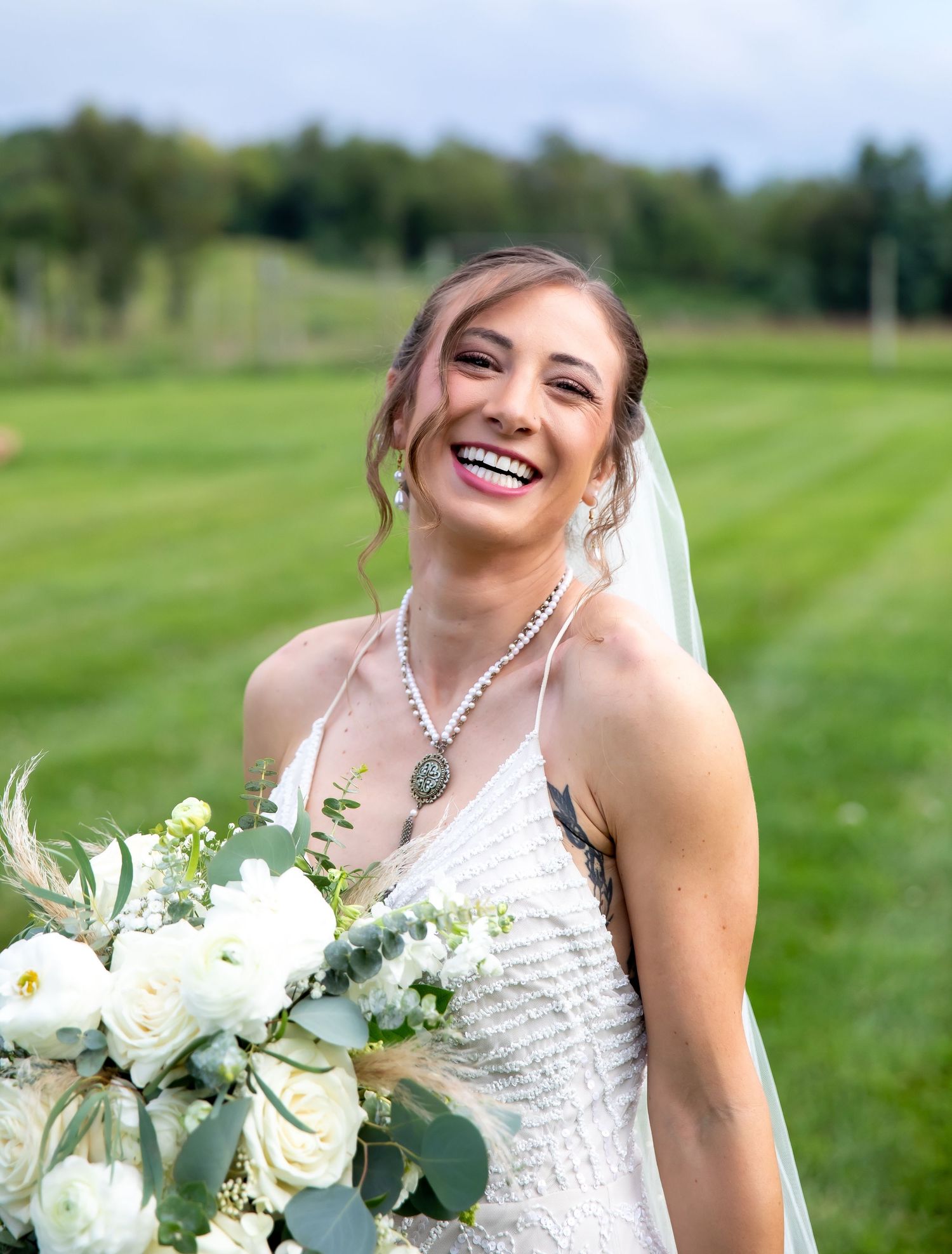 Smiling bride in white beaded gown holding white floral bouquet with eucalyptus, wearing veil and necklace outdoors.