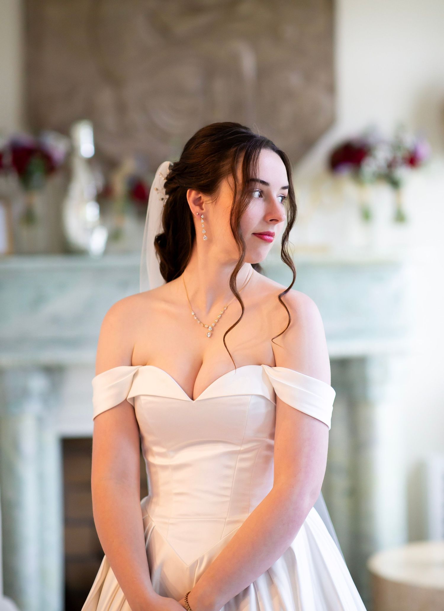 Elegant bride in off-shoulder white satin gown with veil, posing near a fireplace adorned with red floral arrangements.