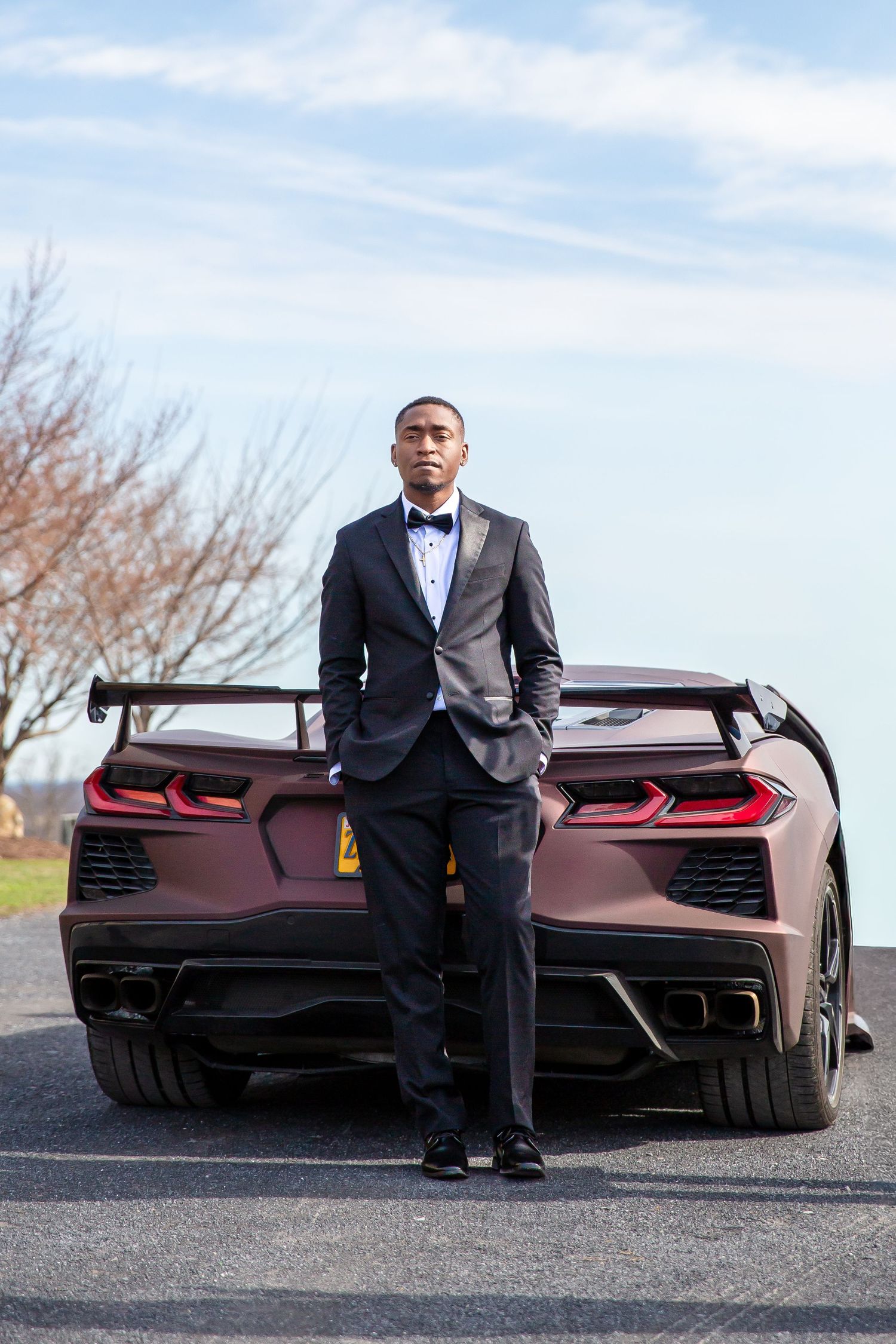 Young Black man in black tuxedo standing in front of sports car outdoors.