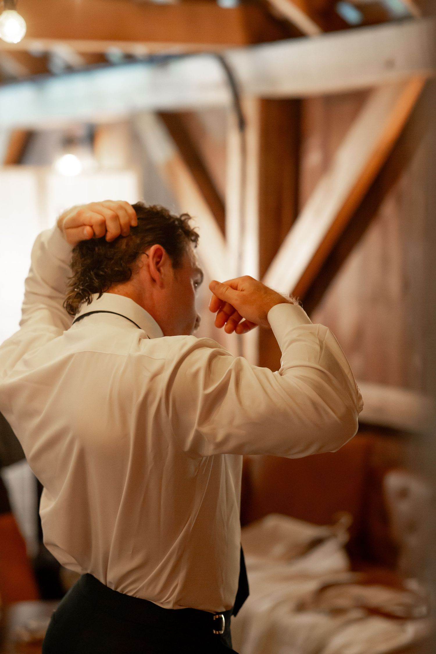 A groom in a white dress shirt adjusts his hair while getting ready in a rustic barn venue before his wedding.