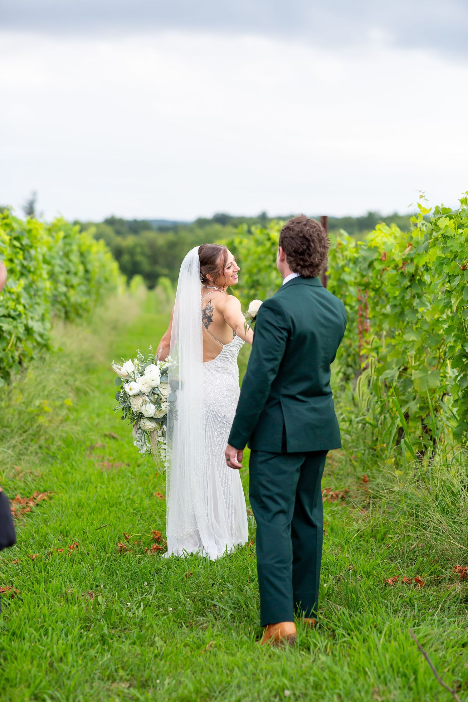 Bride and groom walk hand in hand through lush vineyard rows, bride wearing white gown with veil and bouquet.