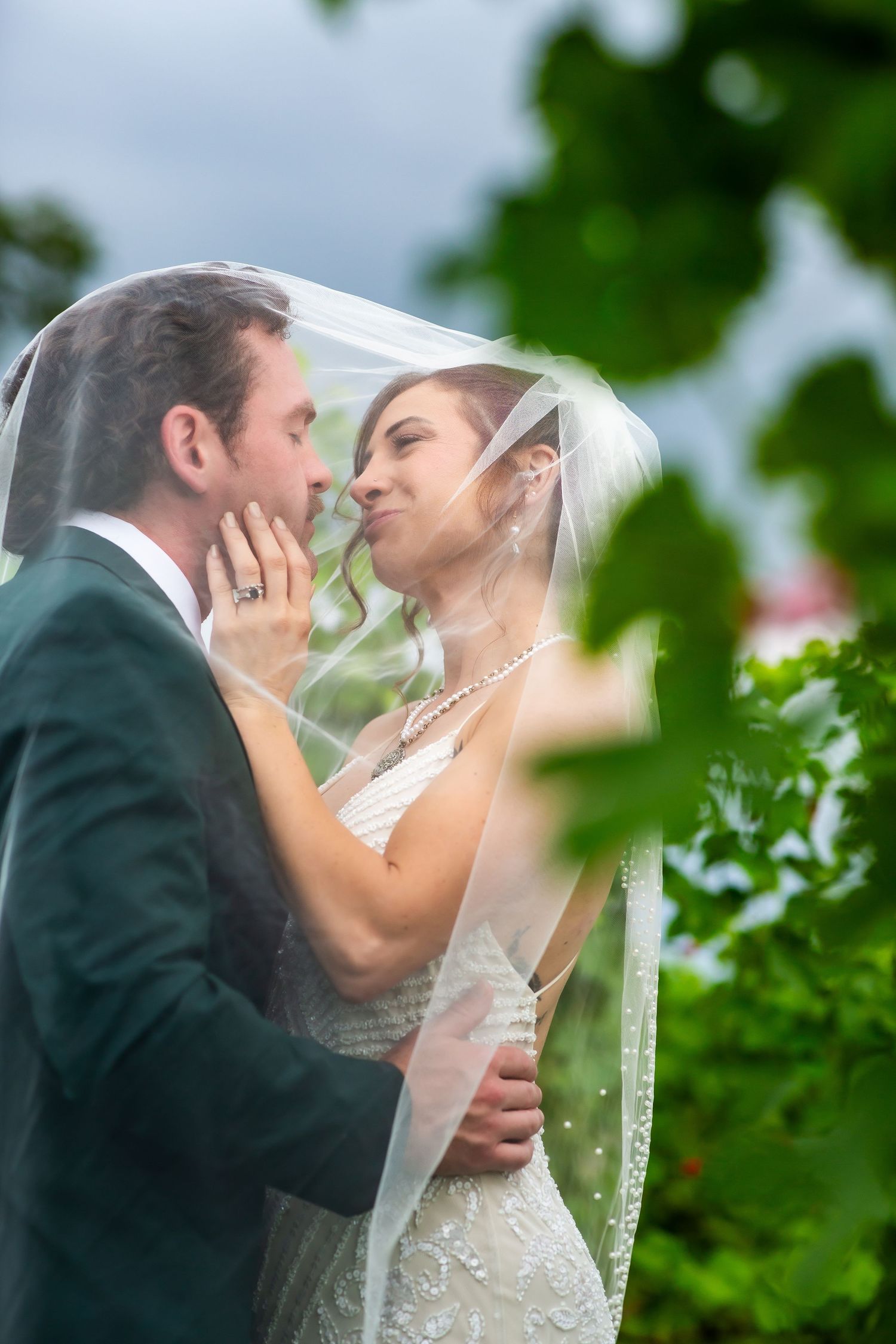 Bride and groom share a romantic moment under a wedding veil outdoors, surrounded by lush green foliage.