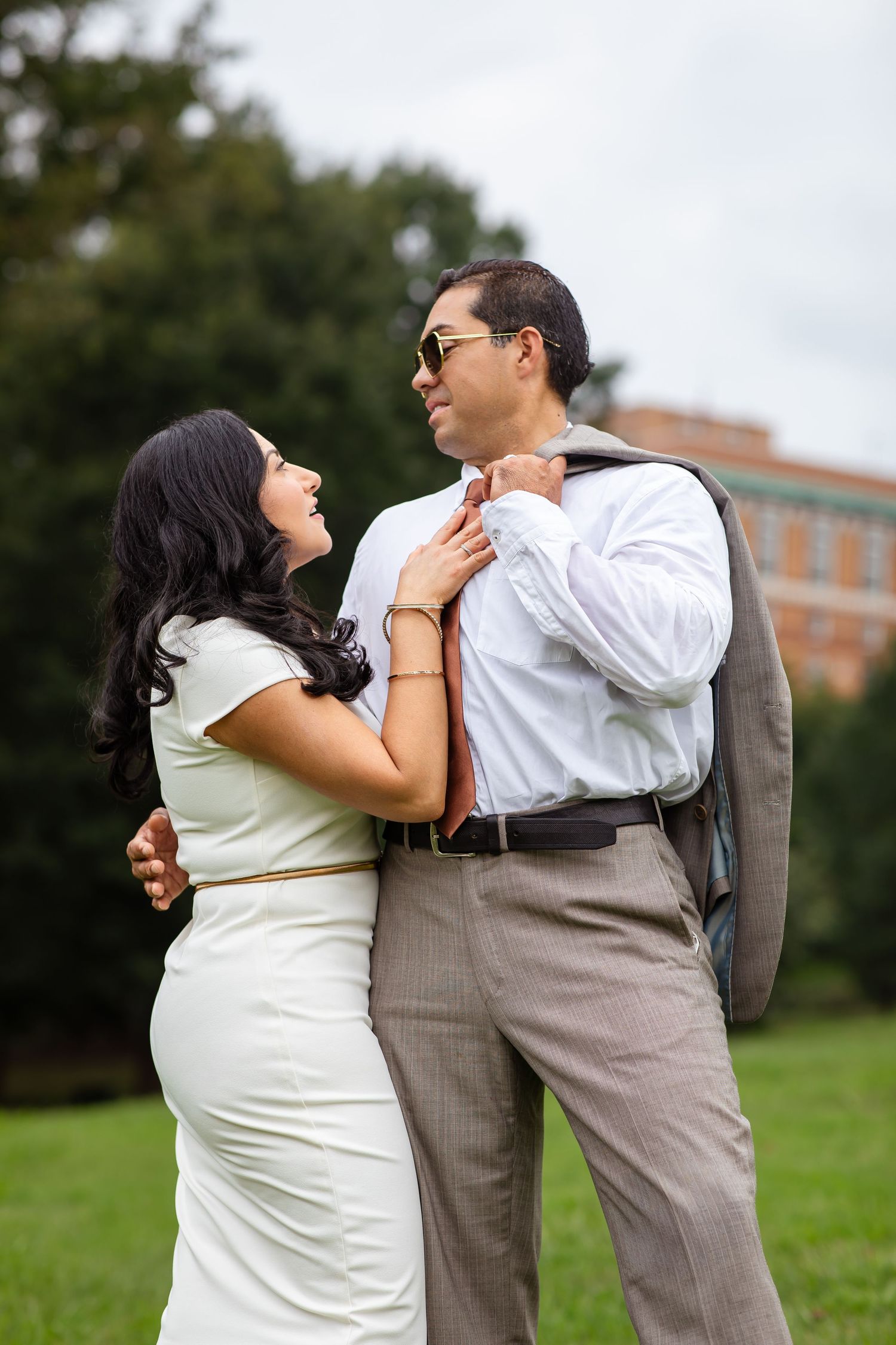 A couple shares a romantic moment outdoors, woman in white dress smiling up at man in sunglasses and gray suit.