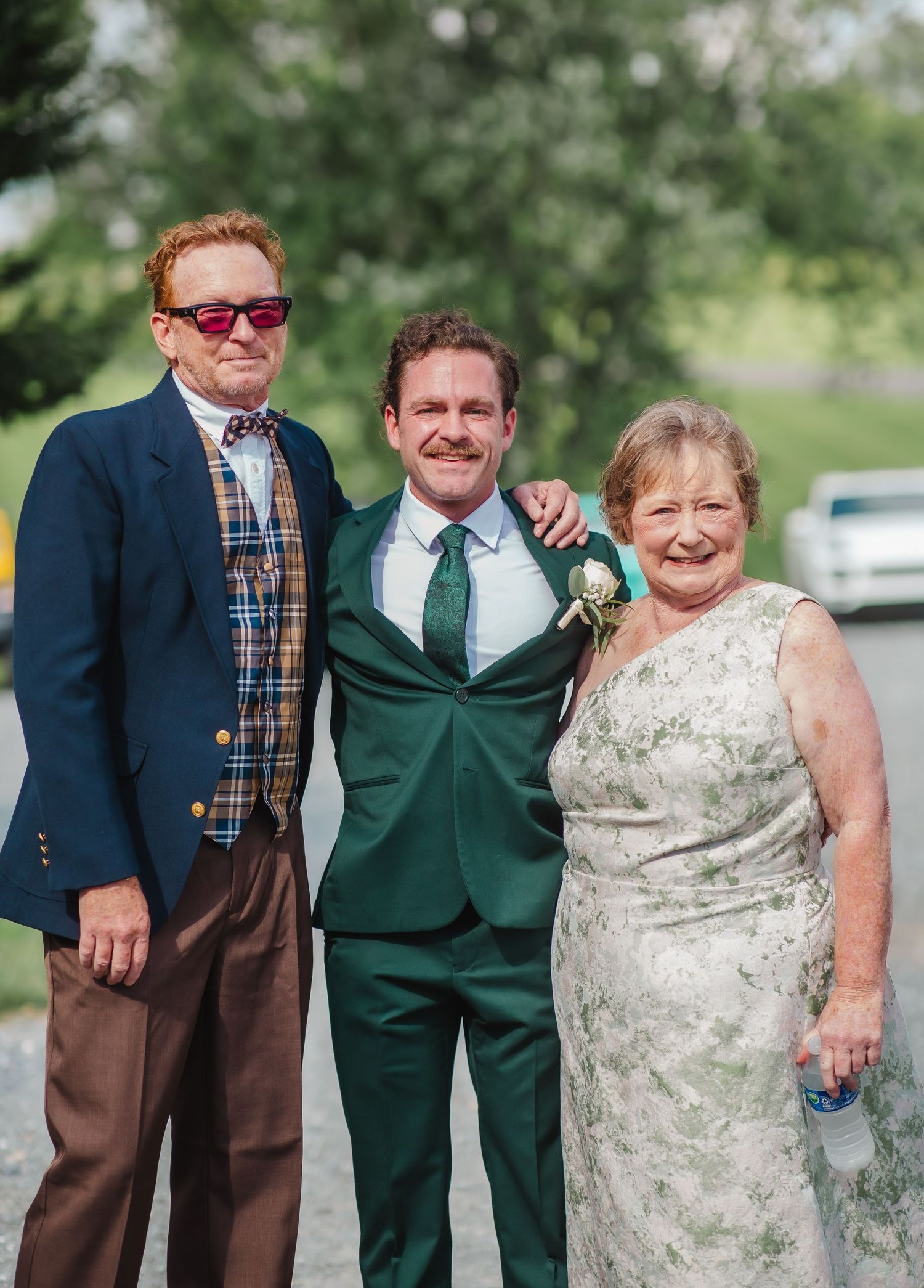 Three people posing outdoors at a wedding, man in green suit with boutonniere flanked by older couple in formal attire.