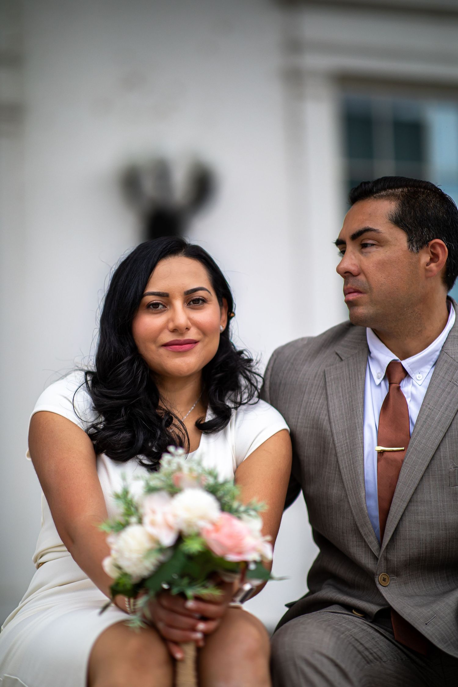 A couple in wedding attire sits together, the bride holding a pink and white bouquet while wearing a white dress.