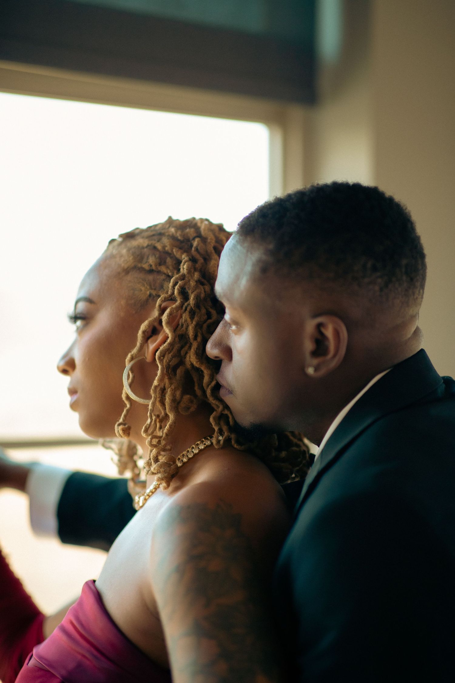 A stylish Black couple in profile looking out a bright window, man in suit, woman in red gown.