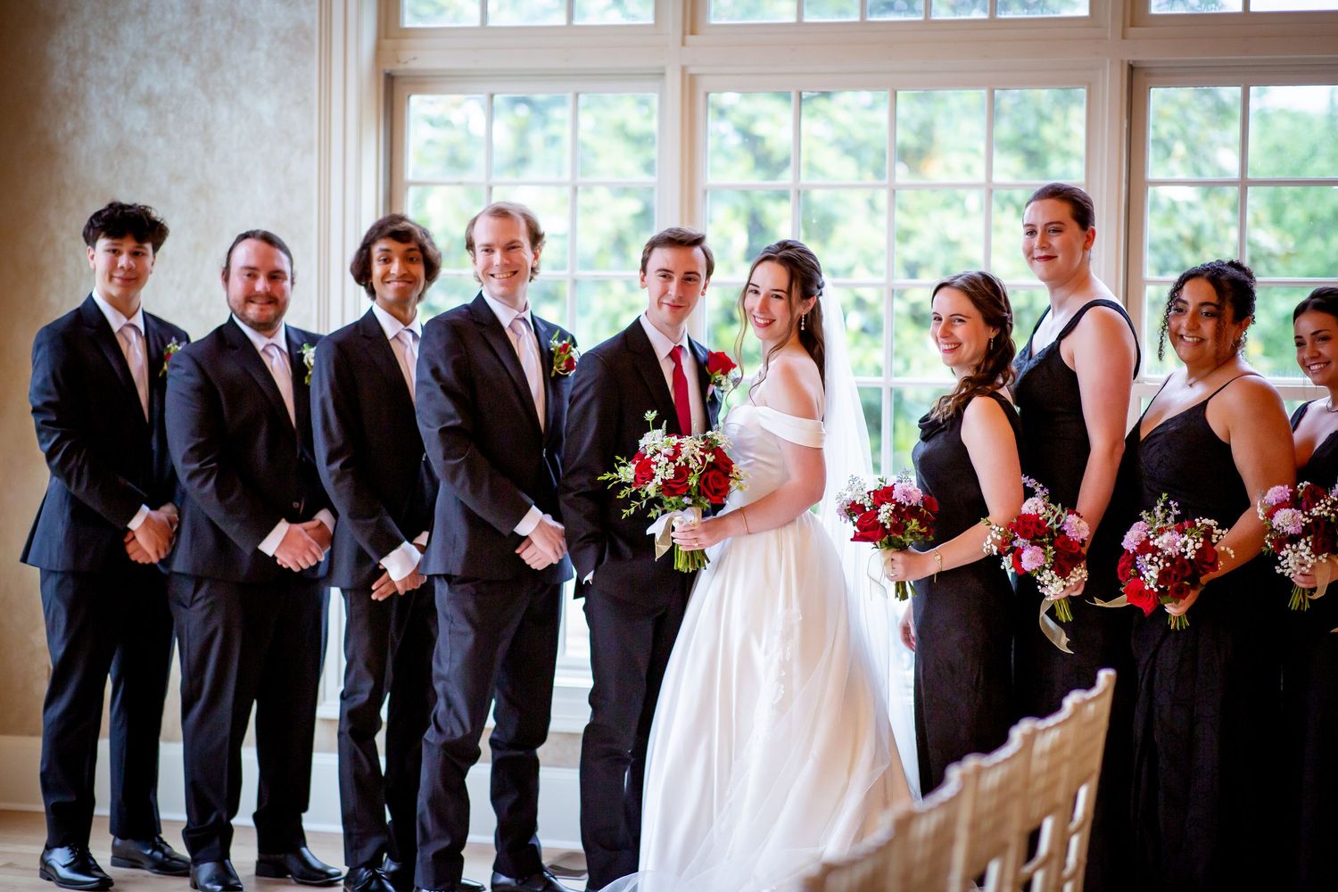 Wedding party poses together, bridesmaids in black gowns and groomsmen in dark suits with floral bouquets.