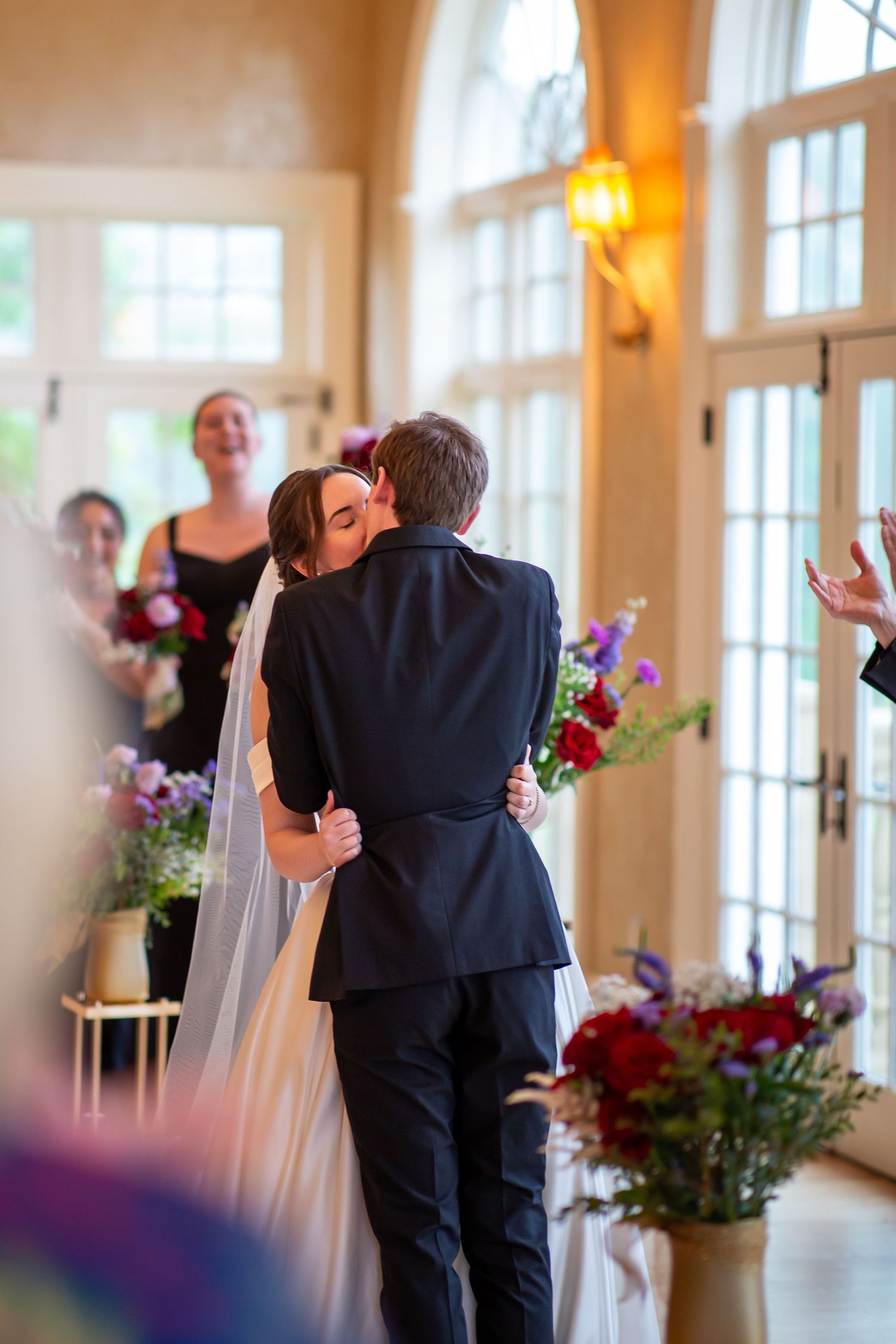 Bride and groom share first kiss at altar surrounded by colorful floral arrangements and cheering guests.