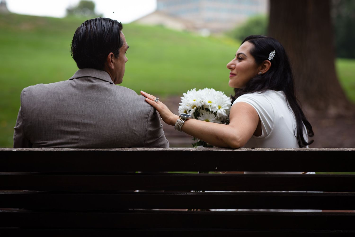 Bride and groom sitting on a park bench, sharing a romantic moment with a white bouquet on their wedding day.