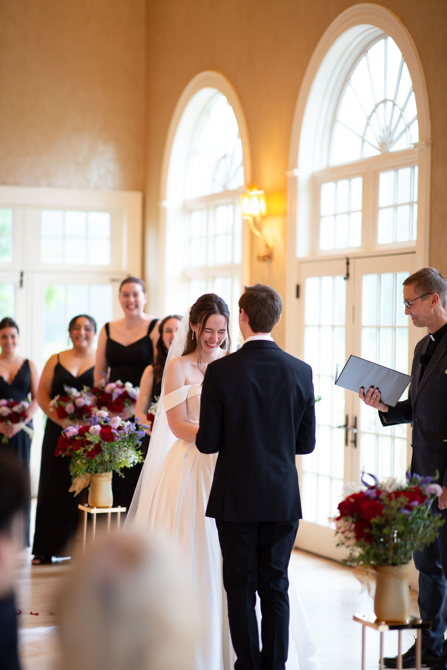 A laughing bride faces her groom at the altar as the officiant reads vows in an elegant venue.
