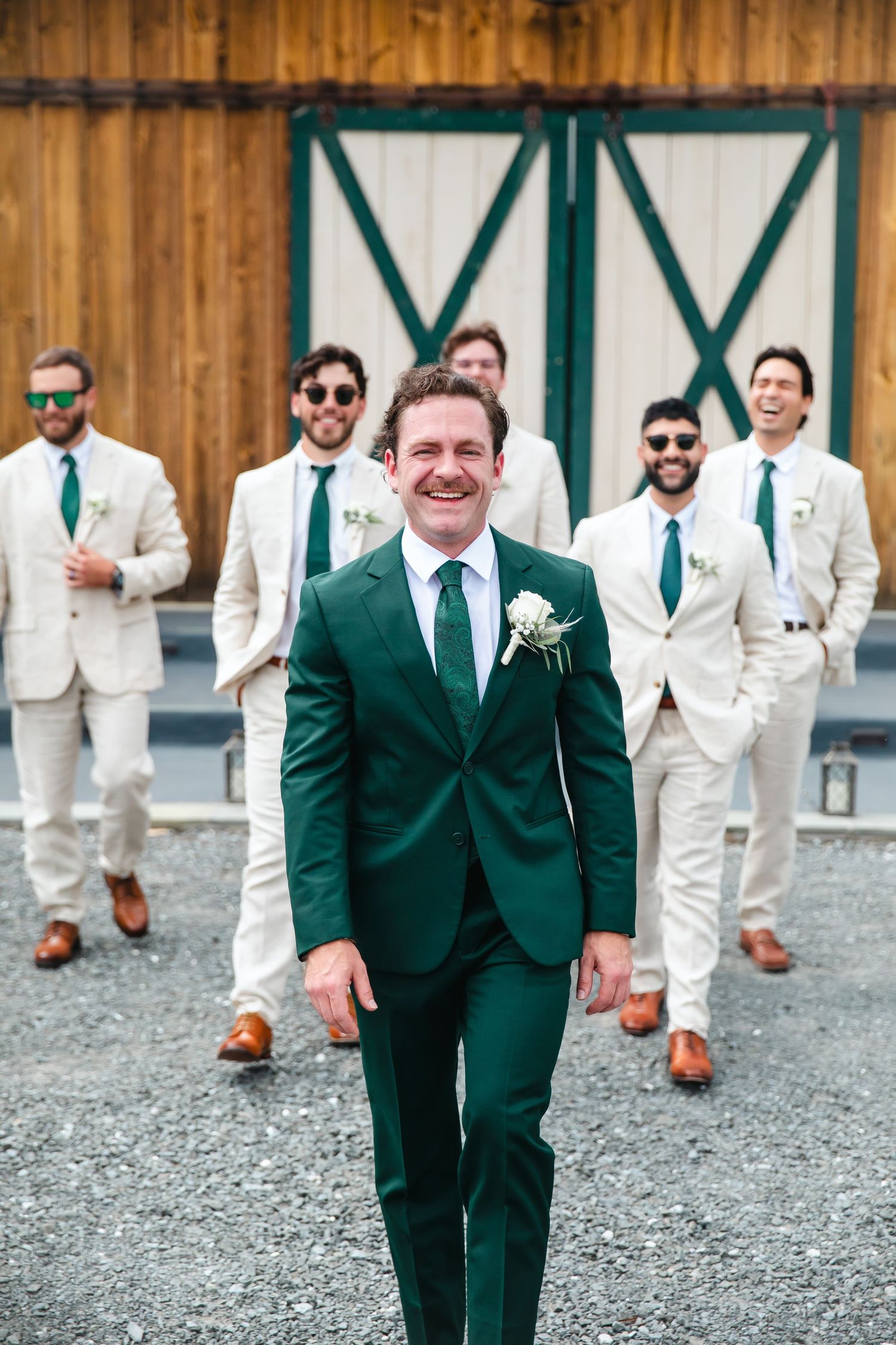 Groom in green suit leads groomsmen in ivory suits with green ties walking outdoors near a barn.