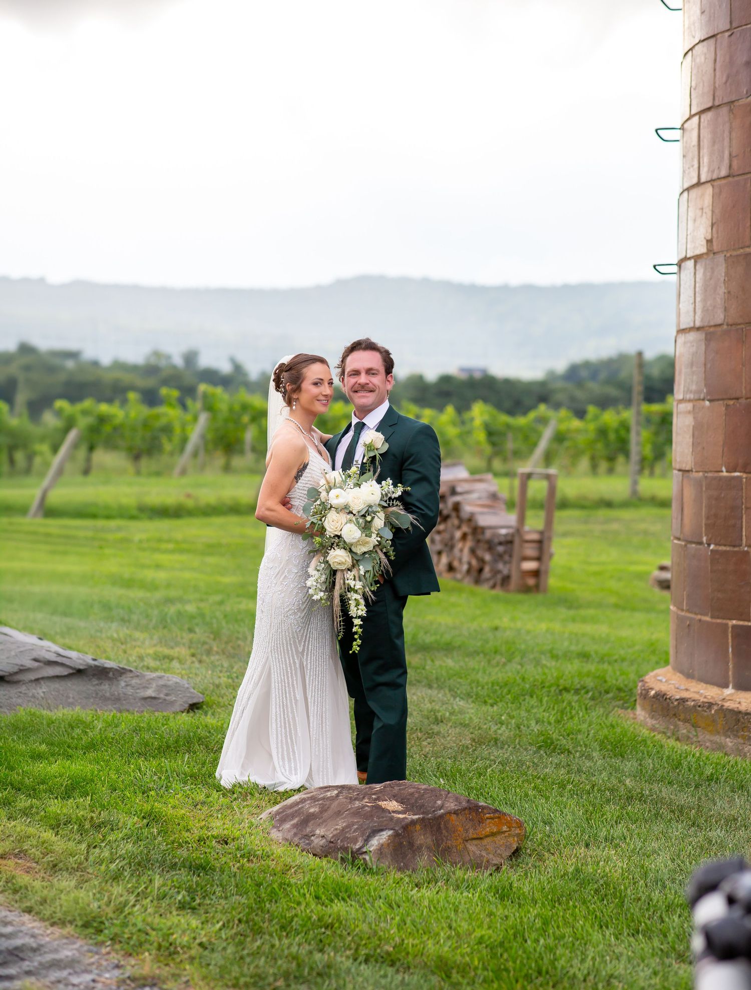 Bride and groom pose in front of lush vineyard with mountain views on their wedding day.