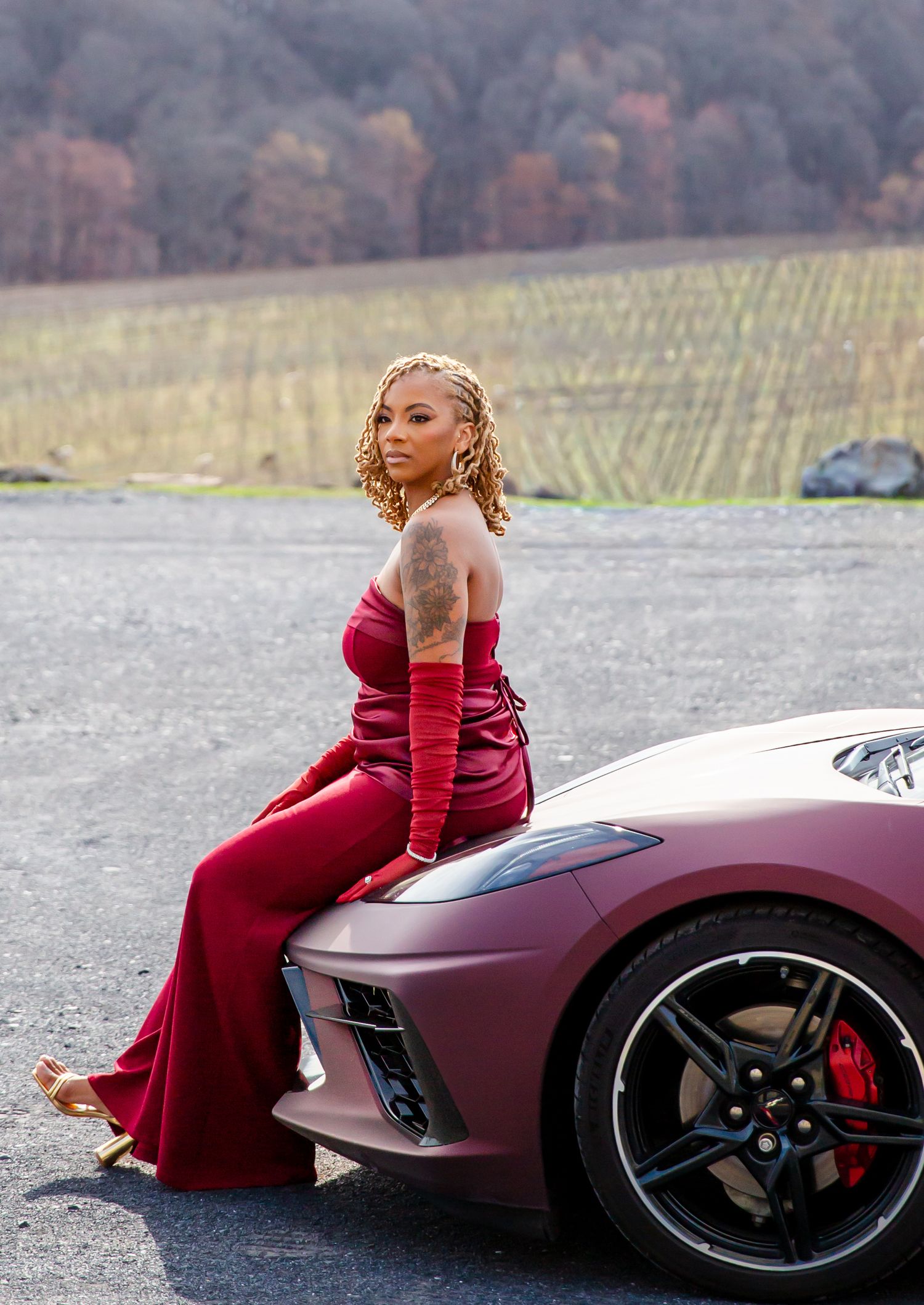 Woman in red strapless gown posing on sports car in scenic setting.