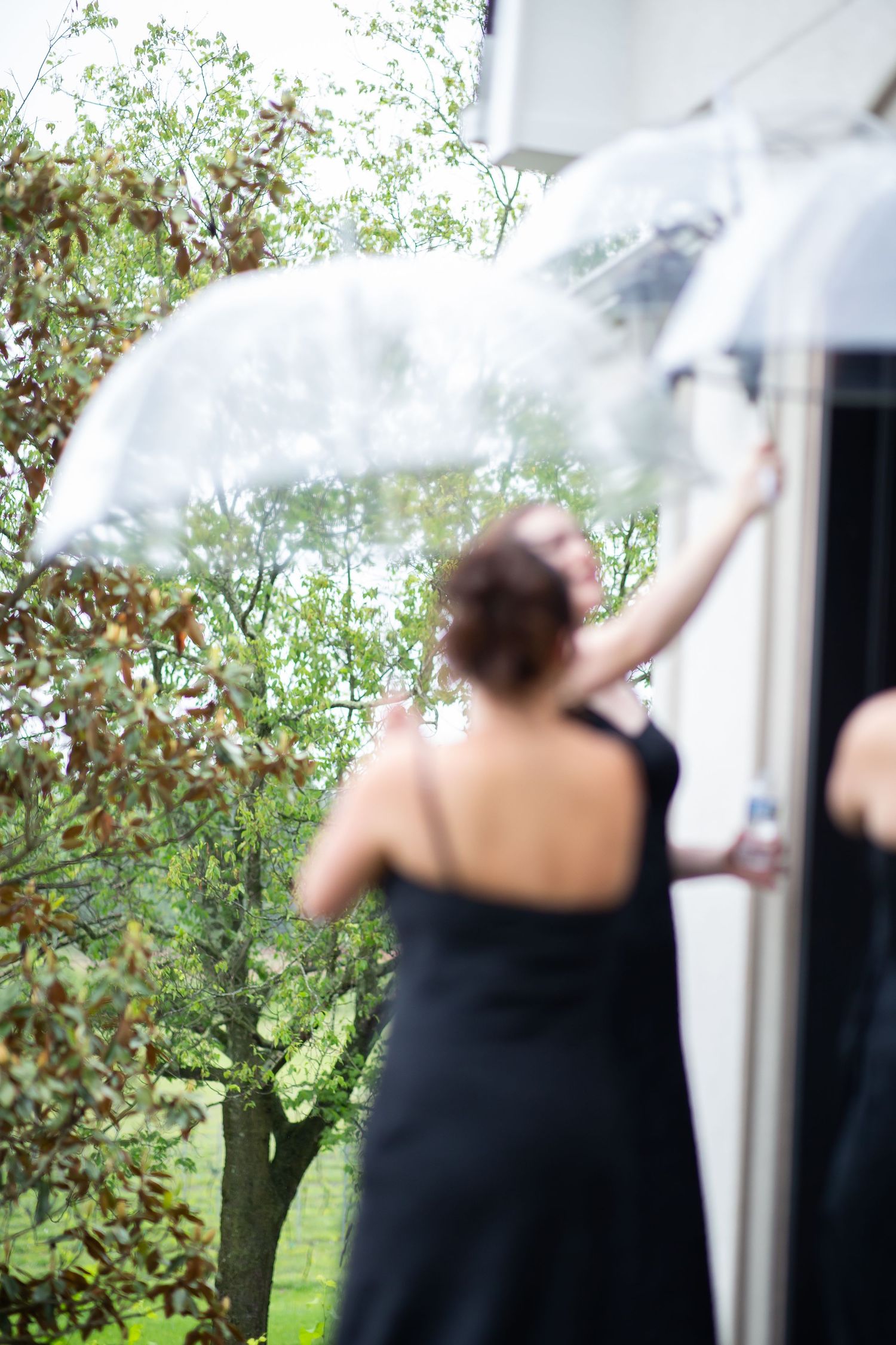 Bridesmaid in black dress holding clear umbrella near white building with trees in background on rainy day.