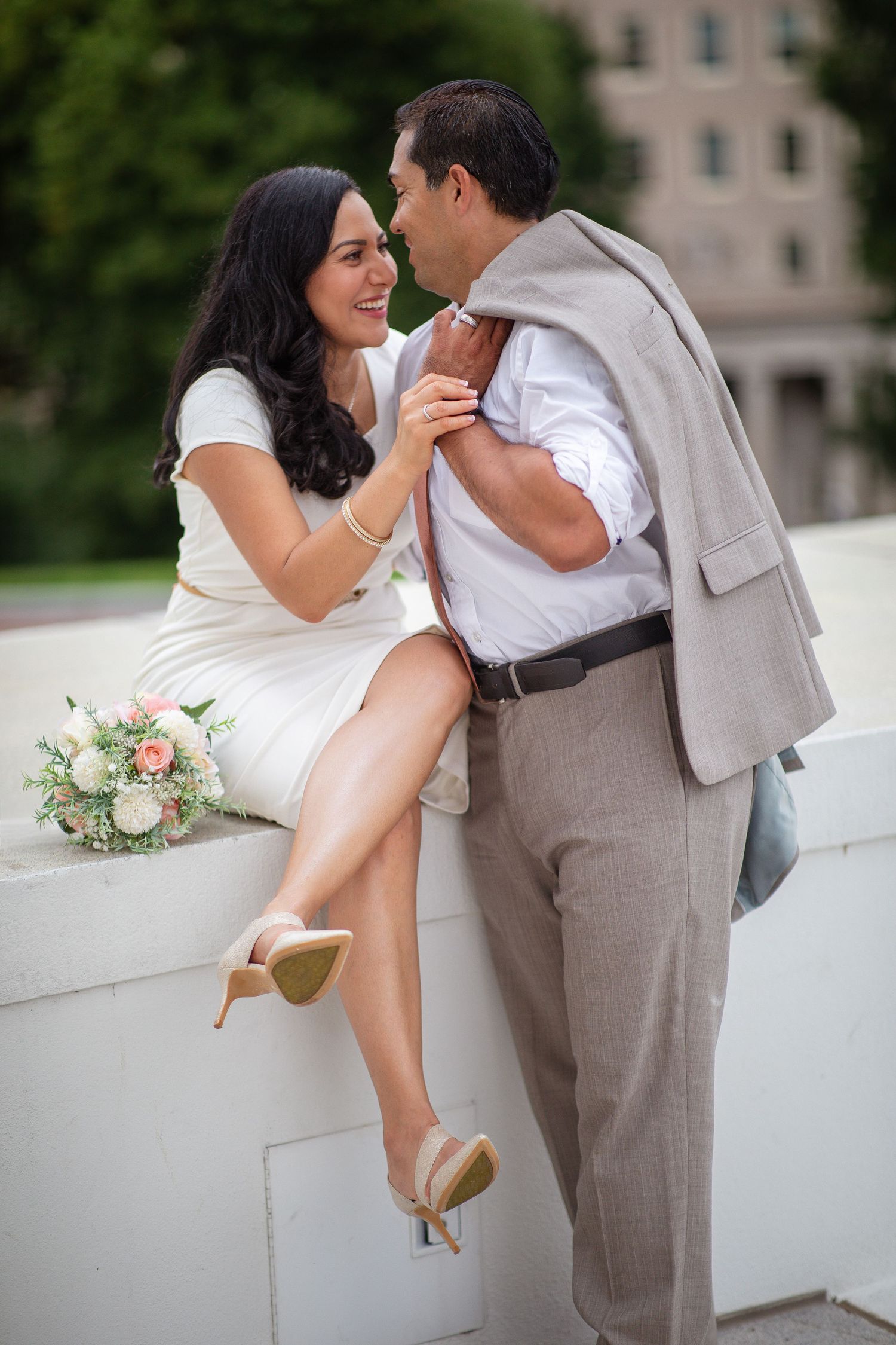 Bride in white dress and groom in gray suit share a romantic moment outdoors, with a pink and white bouquet nearby.