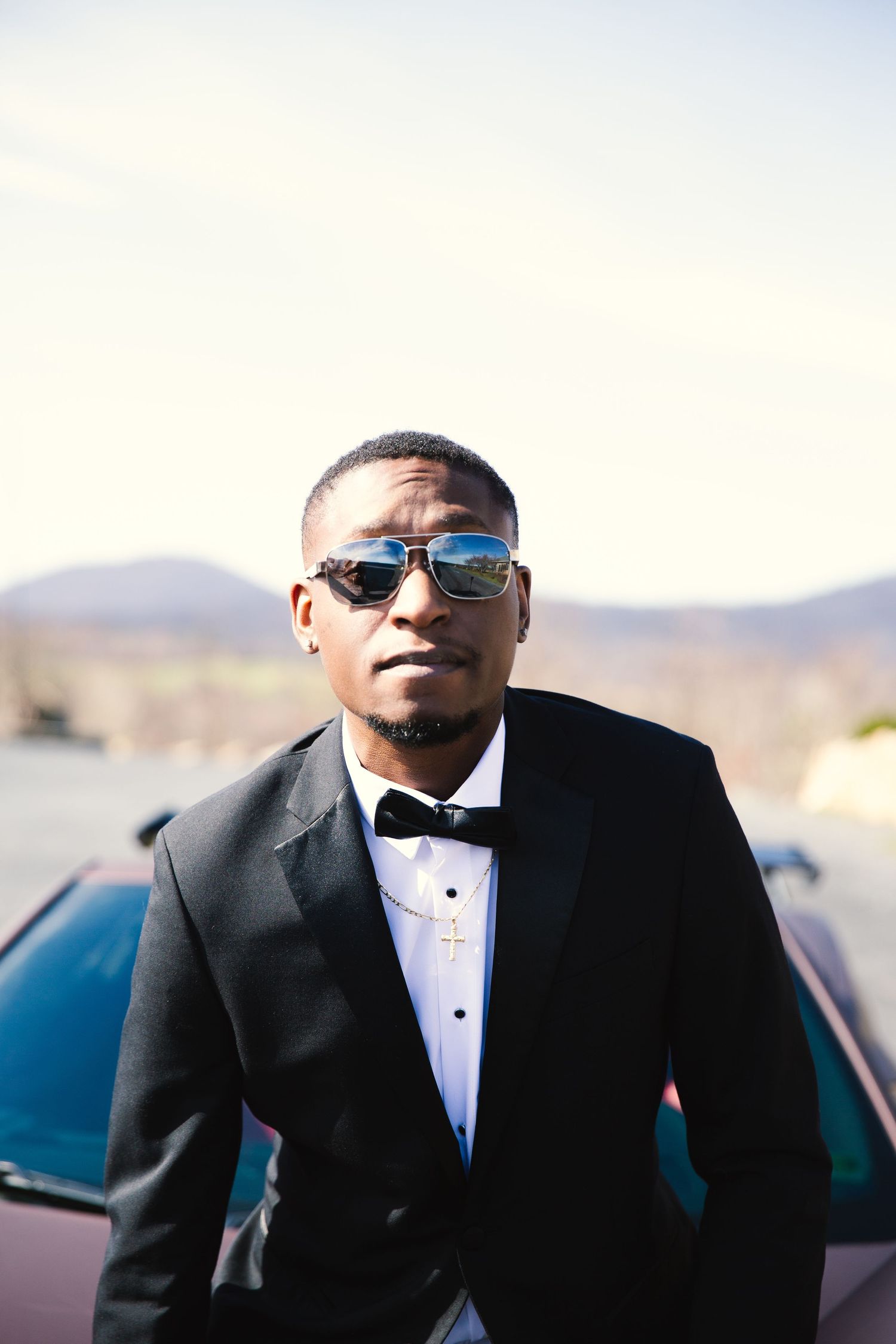 Confident man in black tuxedo with bow tie and sunglasses posing against a red convertible outdoors.