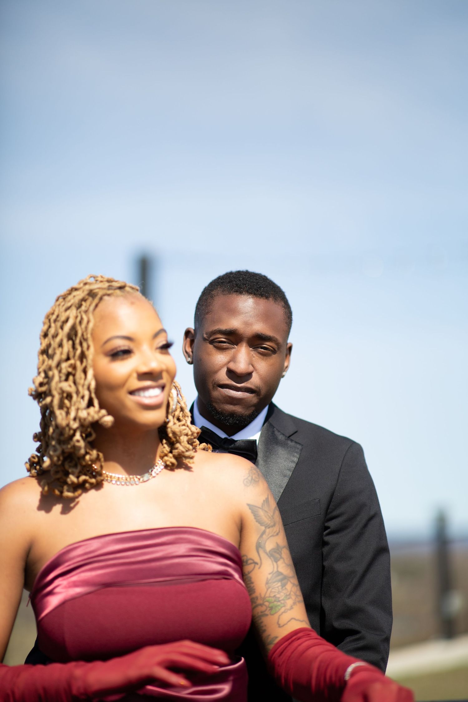 A smiling Black couple outdoors, woman in burgundy strapless dress with red gloves, man in tuxedo.