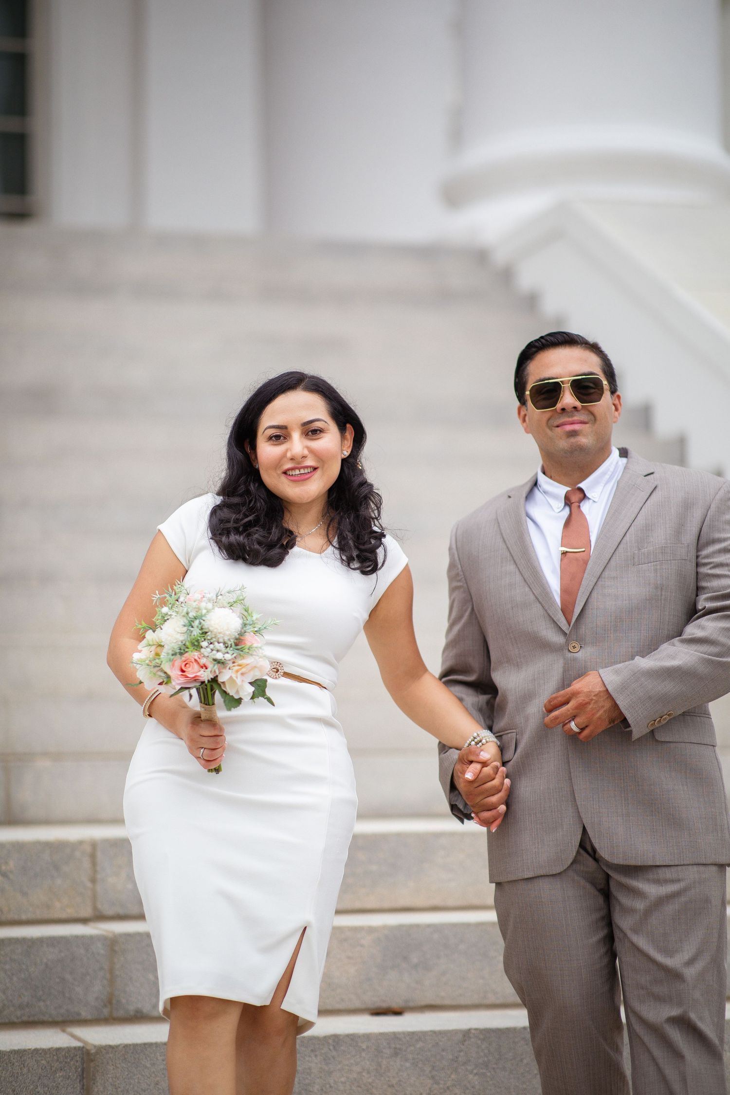 A couple in wedding attire holds hands on steps, bride in white dress with bouquet, groom in tan suit with sunglasses.
